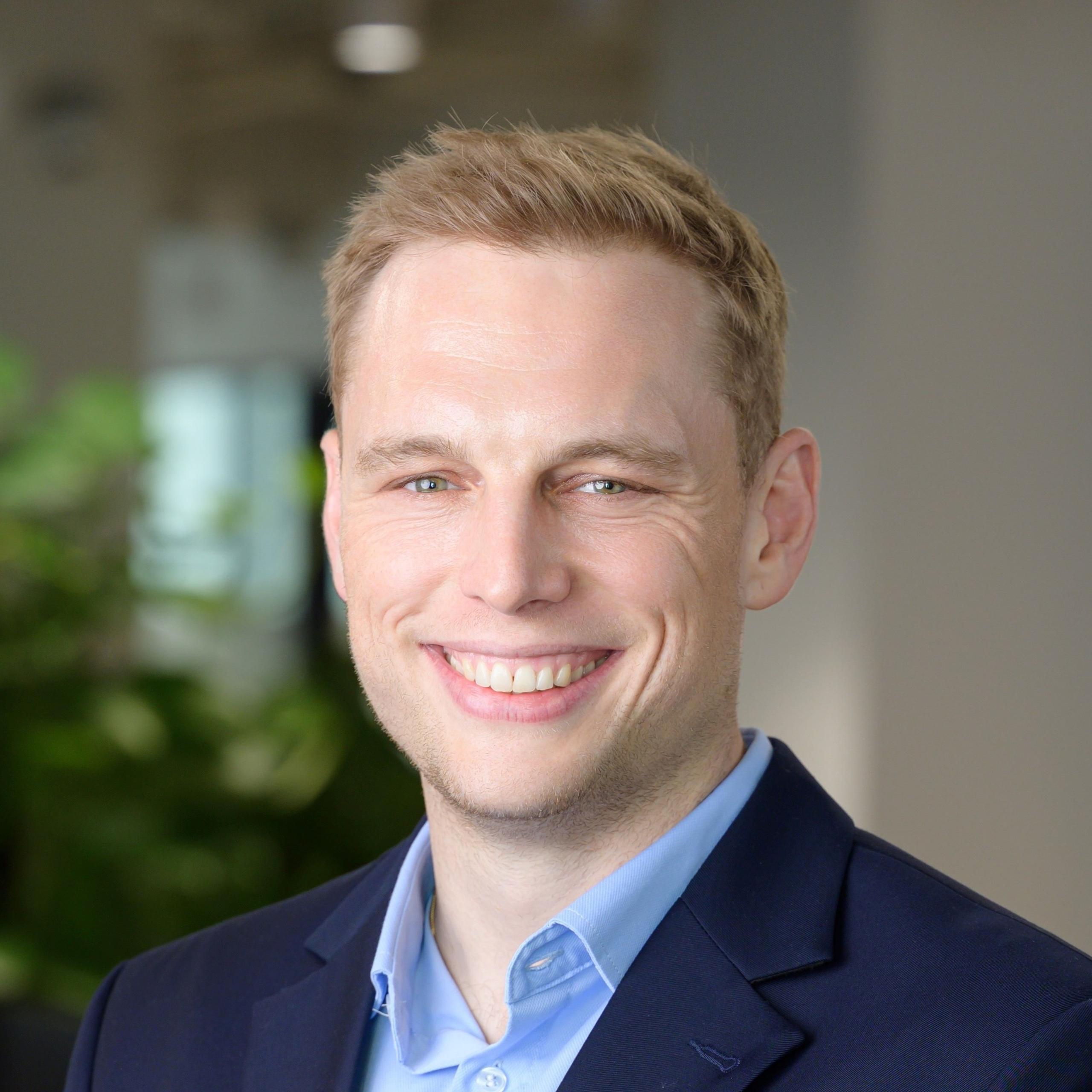 A smiling man in a blue suit stands in a modern office with plants and desks in the background.