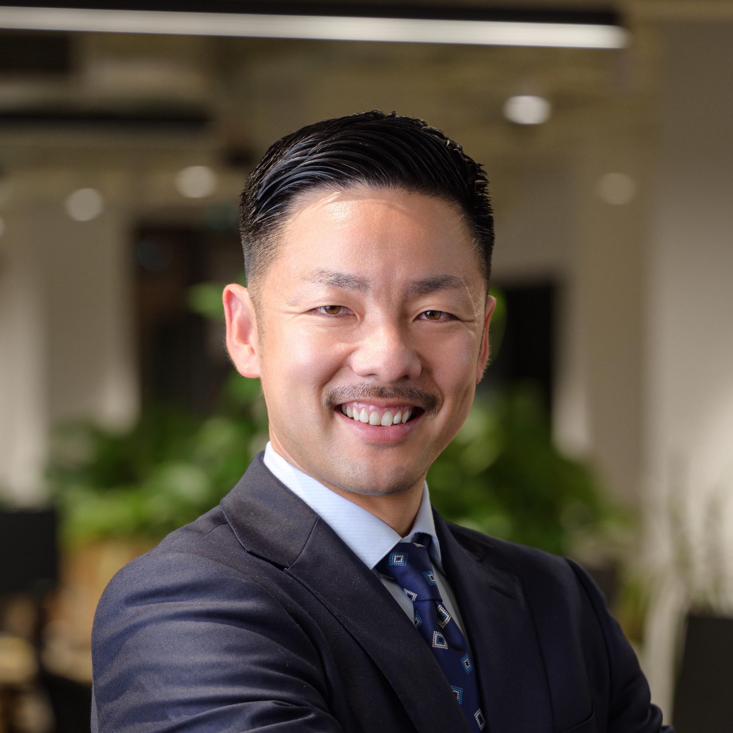 Smiling man in a suit with arms crossed, standing in a modern office with blurred background and plants.