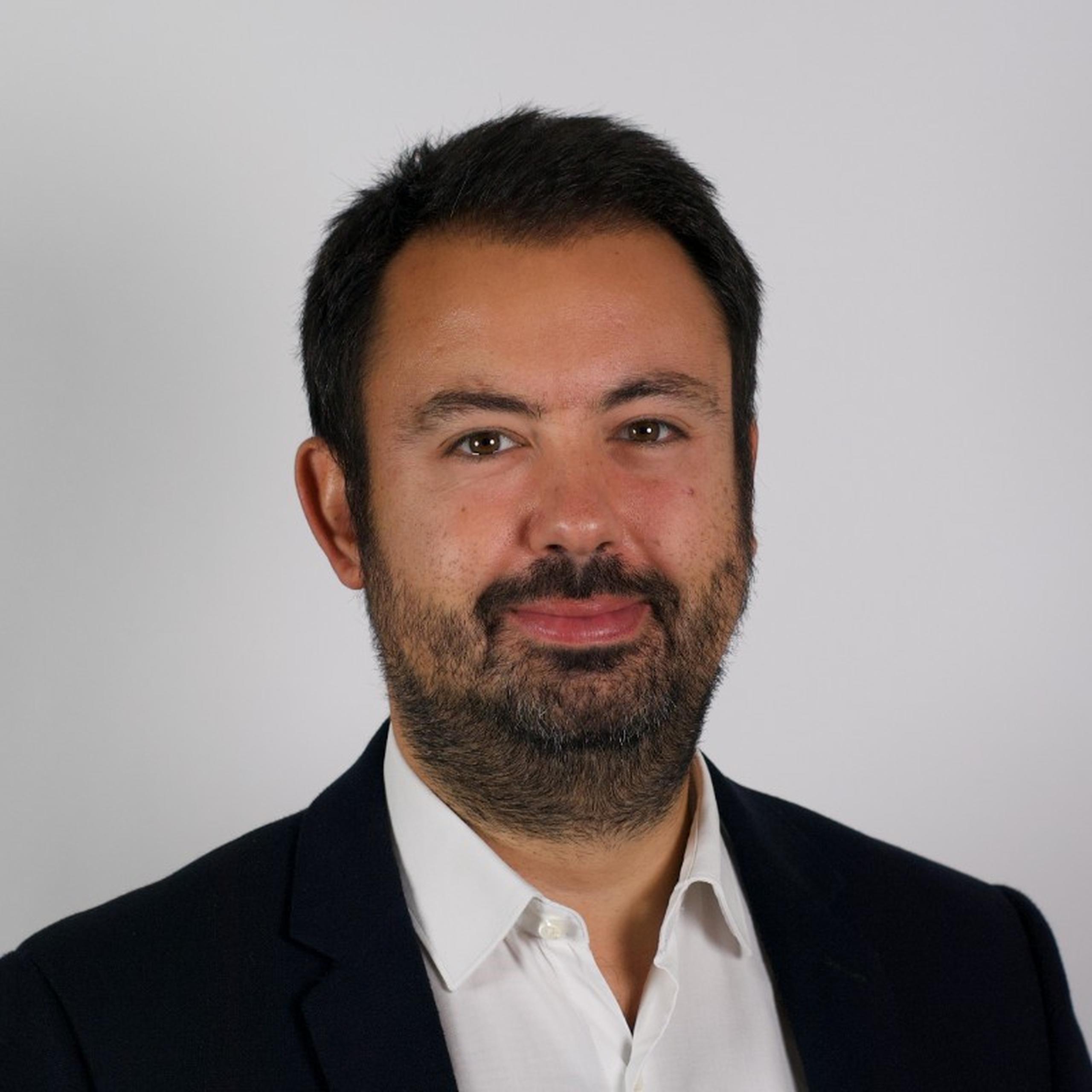 A man with dark hair and a beard, wearing a white shirt and black jacket, smiles slightly against a plain background.