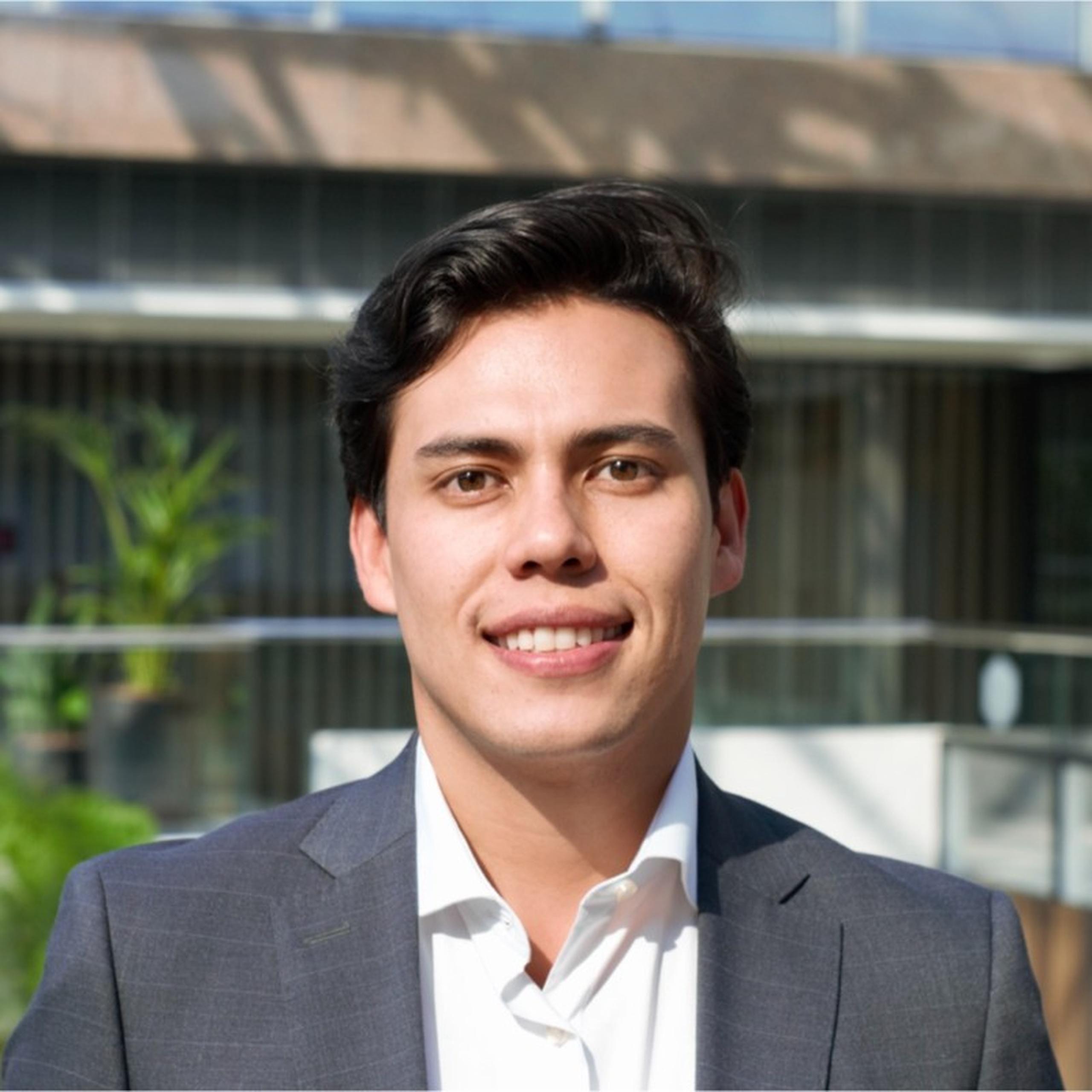 Smiling man in a suit outdoors, standing in front of a modern building with glass panels and greenery.