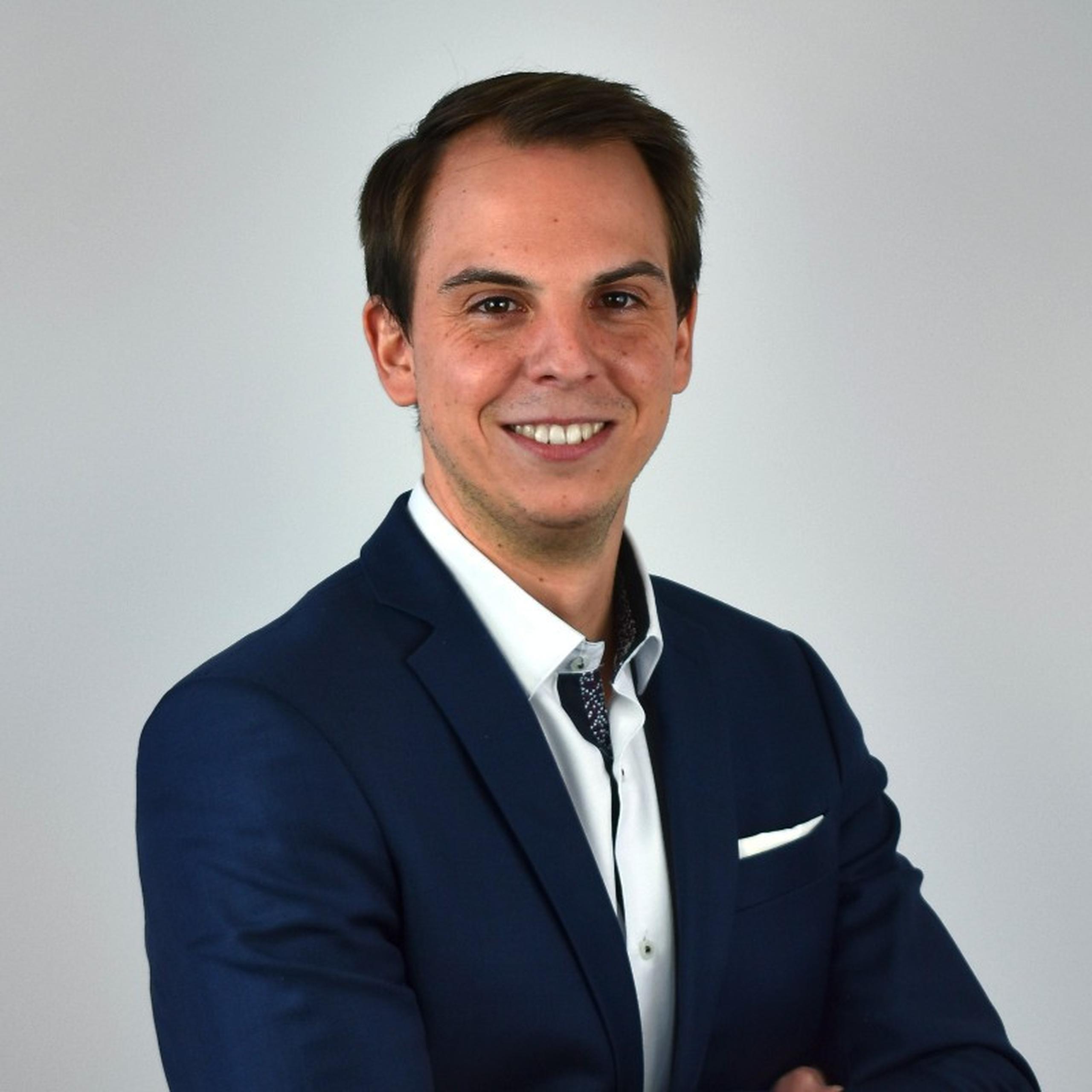 A smiling man in a blue suit and white shirt stands against a plain background, arms crossed confidently.
