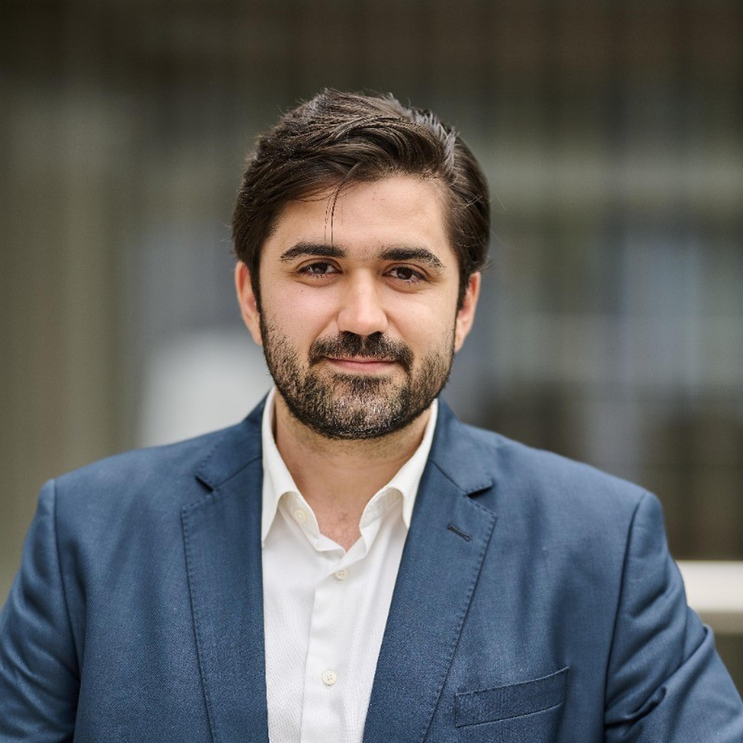 A man with dark hair and a beard, wearing a blue blazer and white shirt, stands outdoors with a neutral expression.