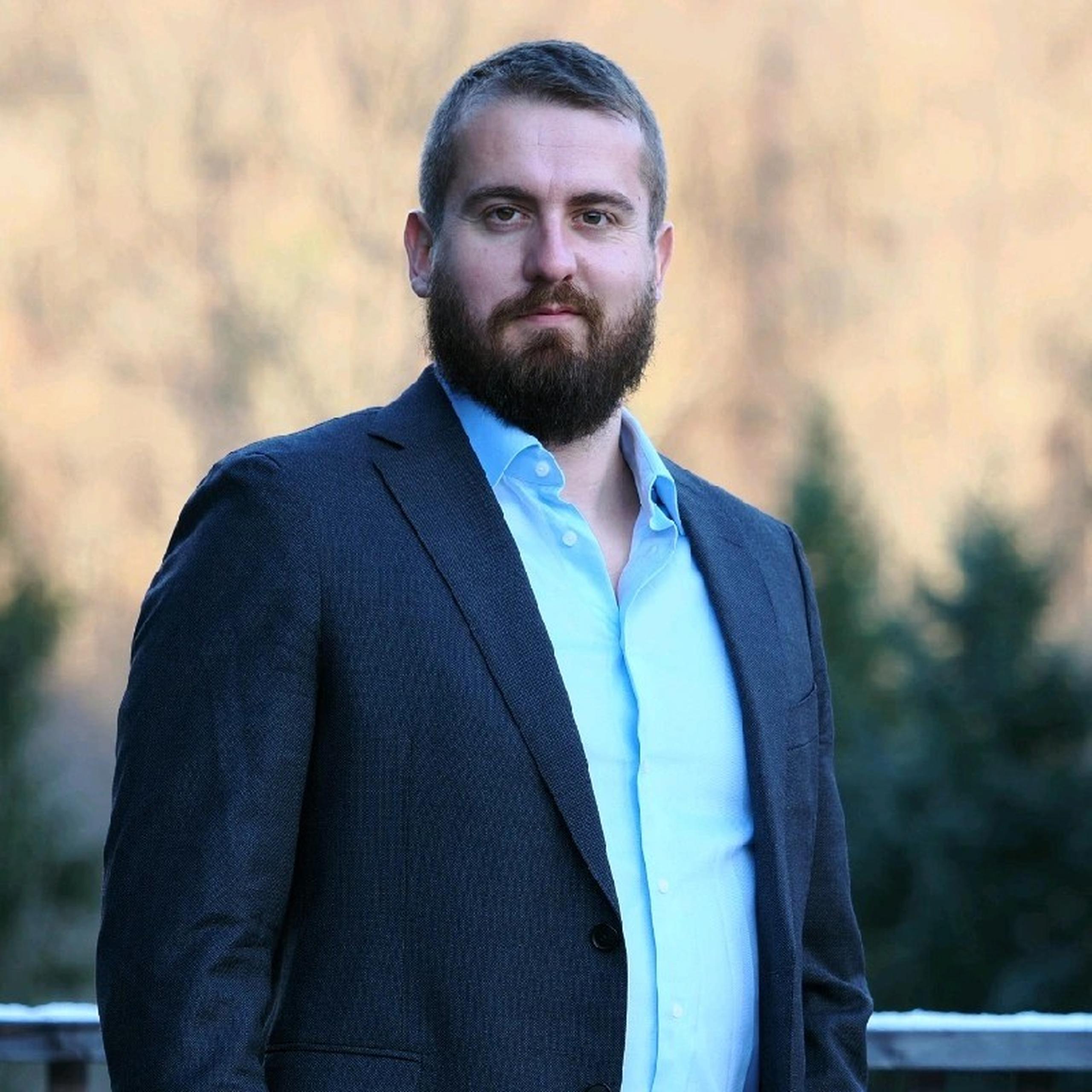 A man with a beard in a suit stands outdoors, with blurred trees in the background.