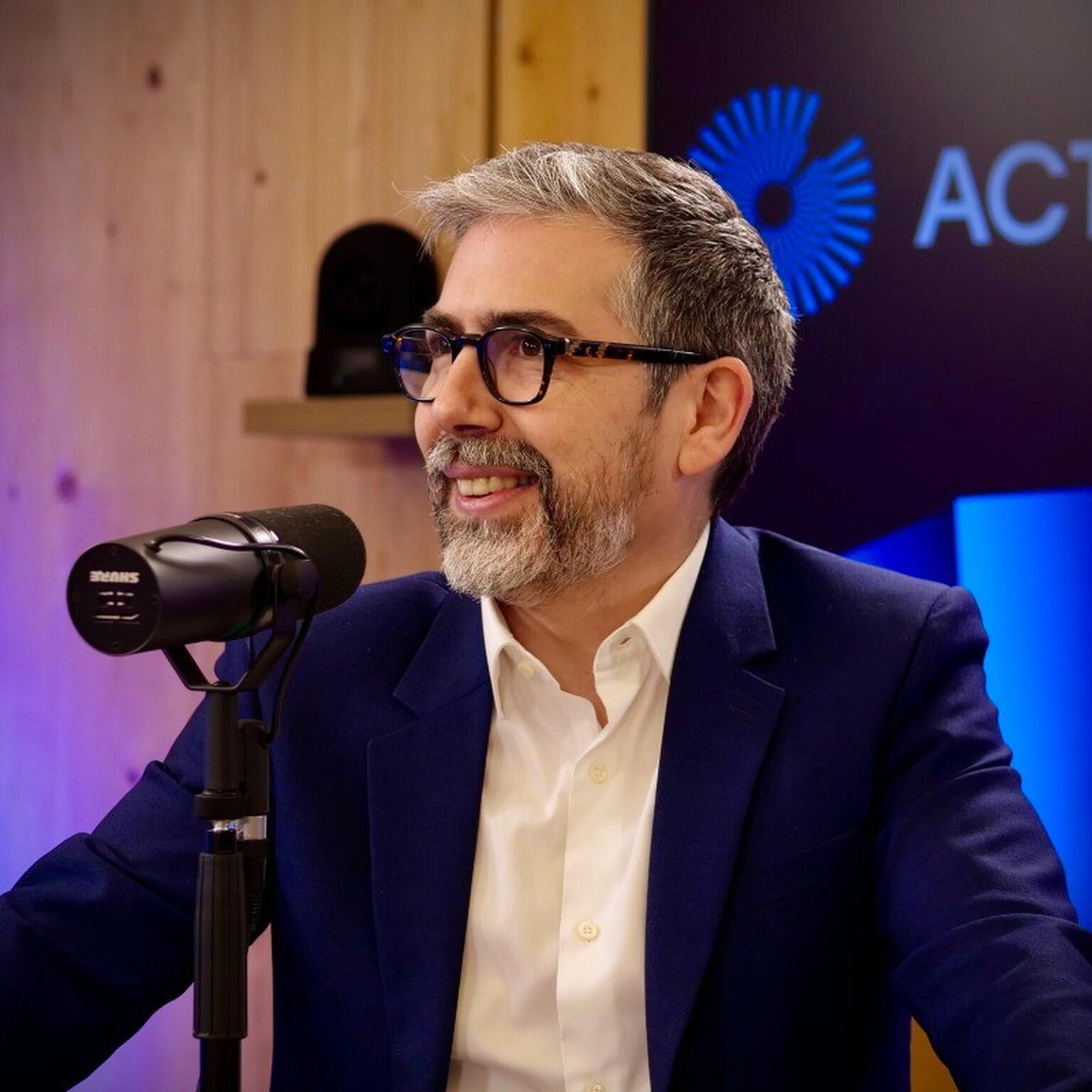 Man with glasses and beard speaking into a microphone in a studio setting, wearing a navy blazer and white shirt.