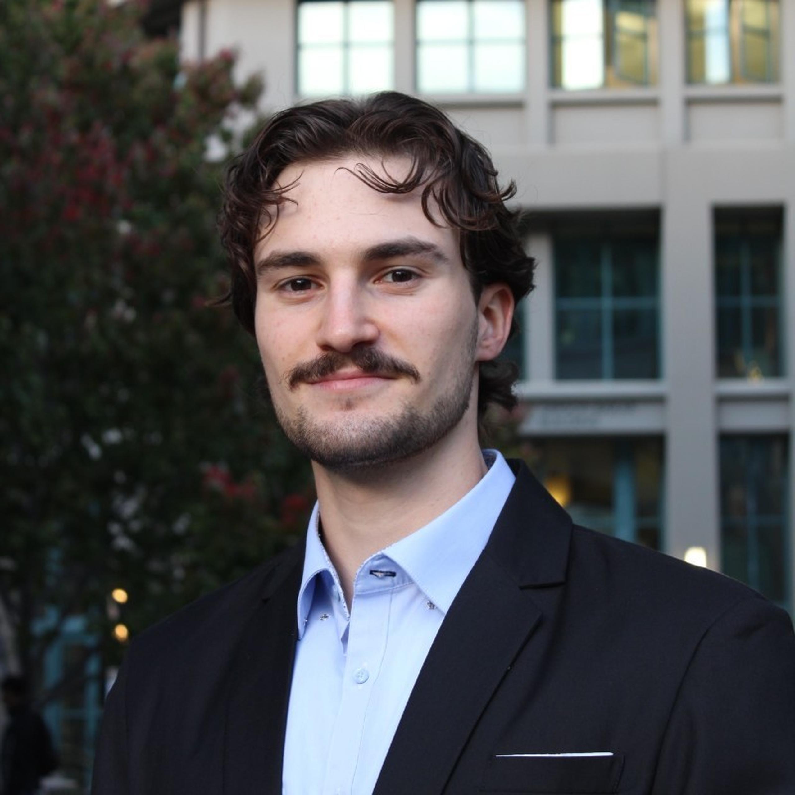 Man with a mustache in a black suit and light blue shirt, standing outdoors in front of a building with large windows.
