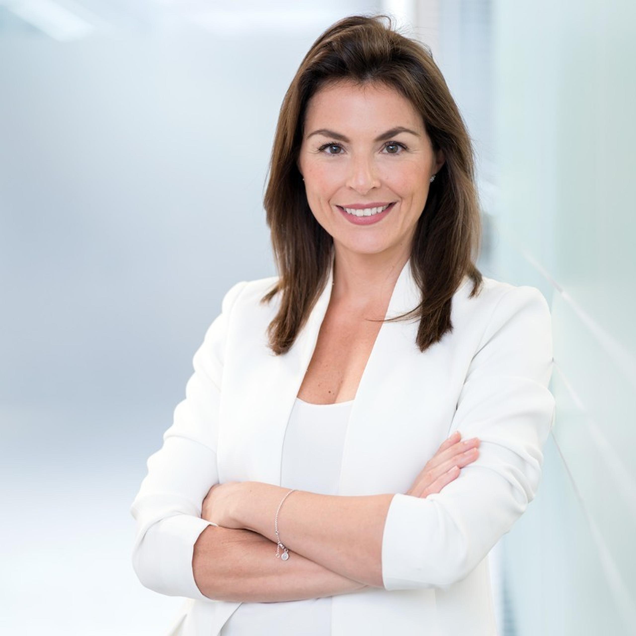 Smiling woman with long brown hair in a white blazer, arms crossed, standing against a blurred light background.