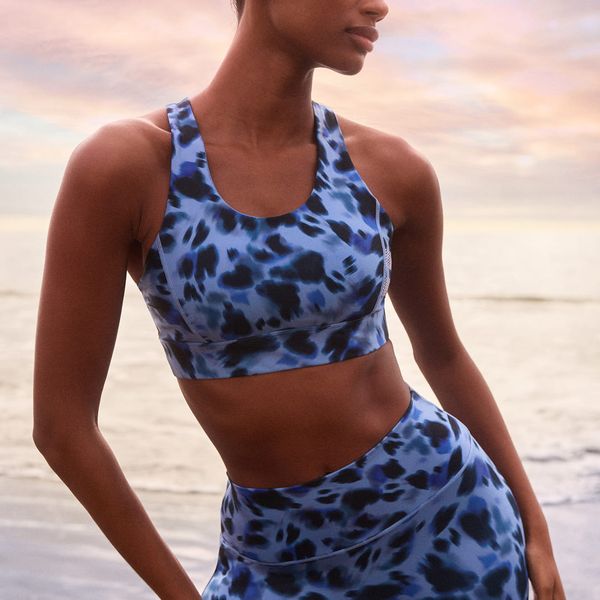 Woman in blue leopard-print activewear poses by the ocean under a colorful sunset sky.