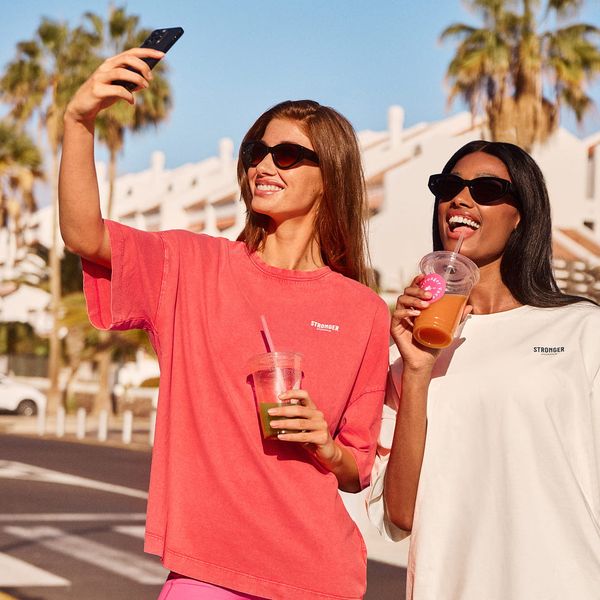 Two women in sunglasses taking a selfie outdoors, holding drinks. They wear oversized shirts, with palm trees and buildings in the background.