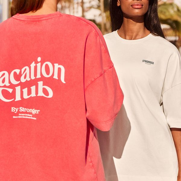 Two women wearing casual t-shirts, one red with "Vacation Club" text and the other white with "STRONGER" logo, stand outdoors in sunlight.