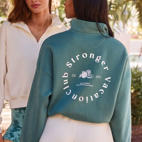 Two women in casual attire; one wearing a green sweater with "Stronger Vacation Club" text on the back, the other in a cream top with a high collar.