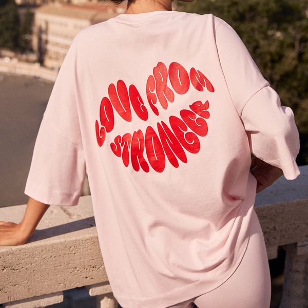 Person wearing a light pink shirt with bold red text "LOVE FROM STRONGER" standing by a stone railing, overlooking a scenic view.