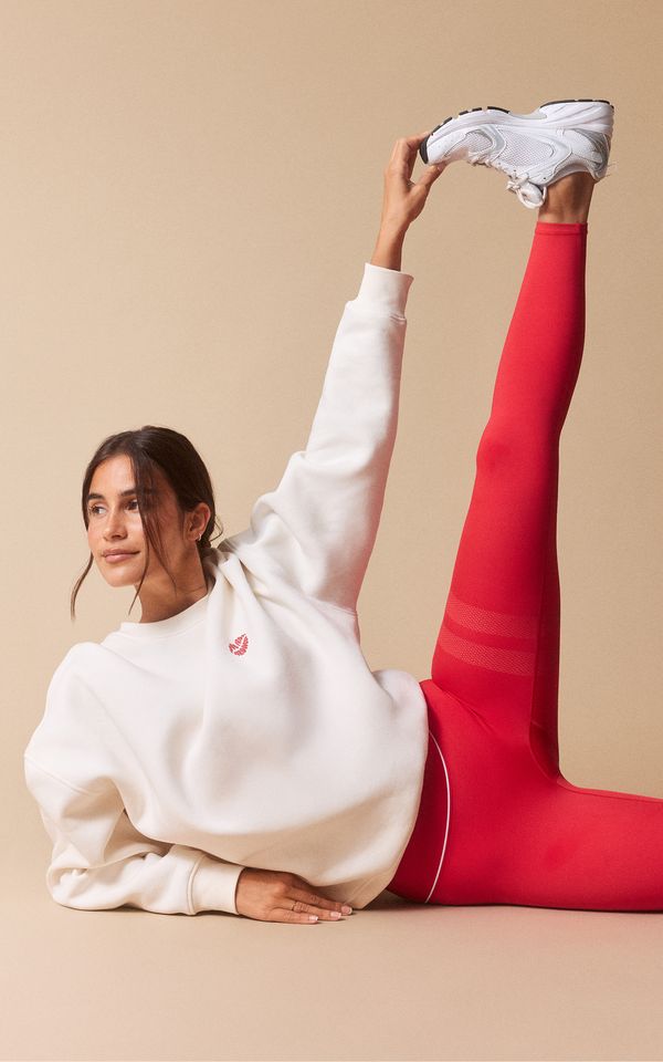 Woman in a white sweatshirt and red leggings performs a leg stretch while lying on the floor, with one leg raised and wearing white sneakers.