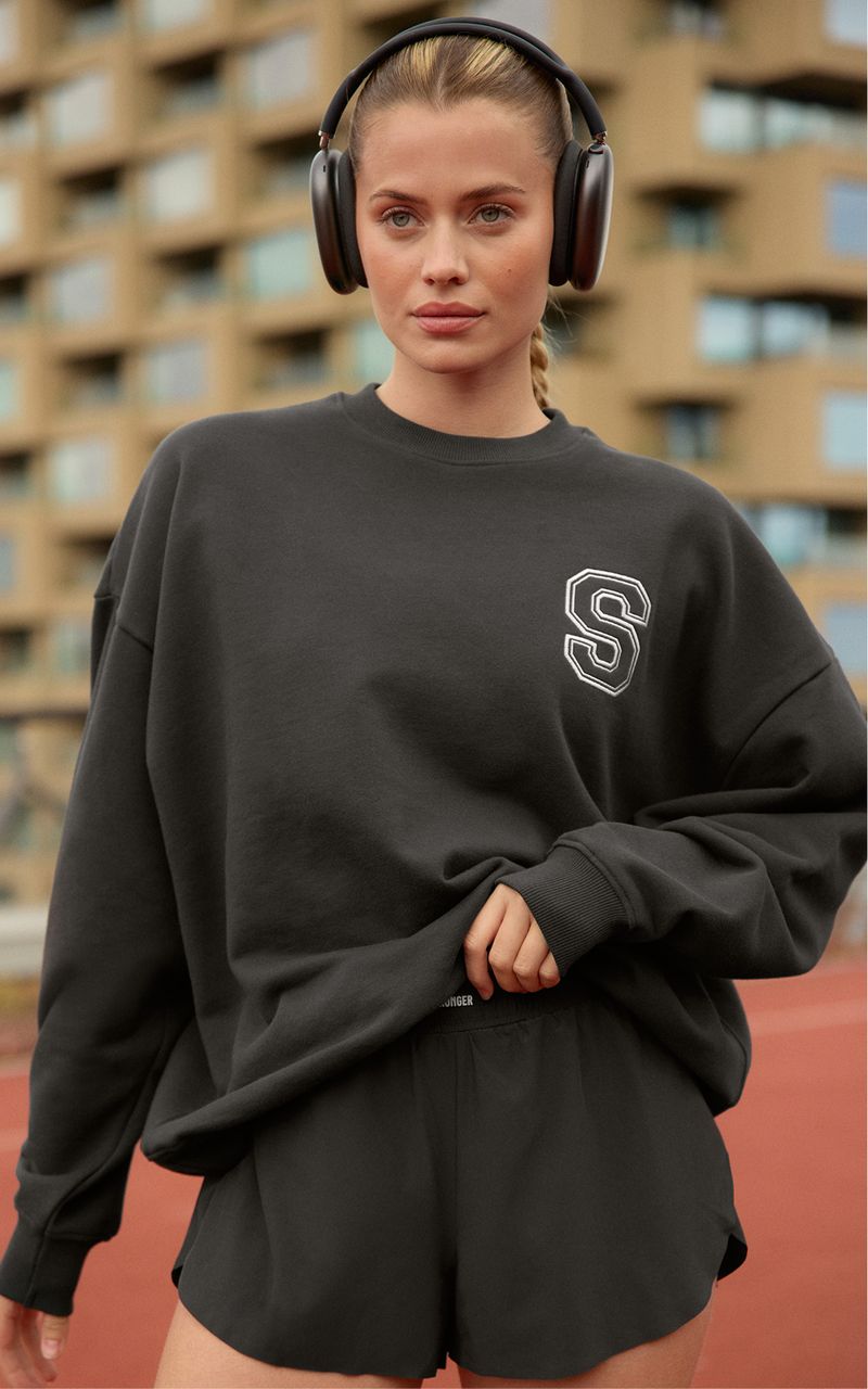 Woman wearing headphones, a dark sweatshirt with a letter "S," and shorts, standing on a track with a building in the background.