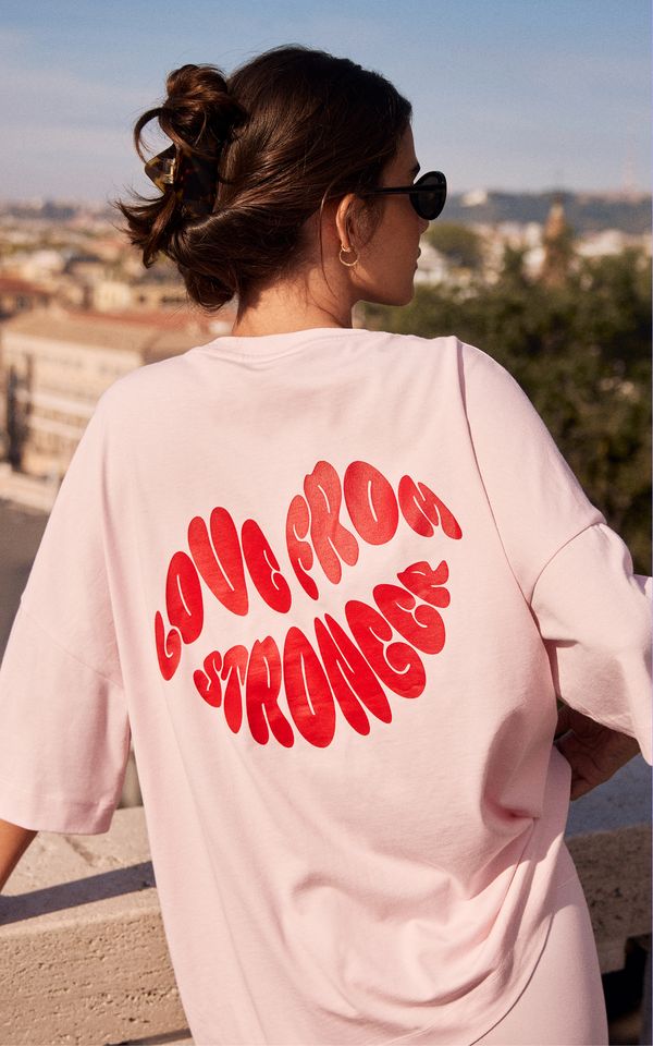 Woman with sunglasses, wearing pink shirt with "LOVE FROM STRONGEST" in red letters, leans on a stone railing overlooking a cityscape.