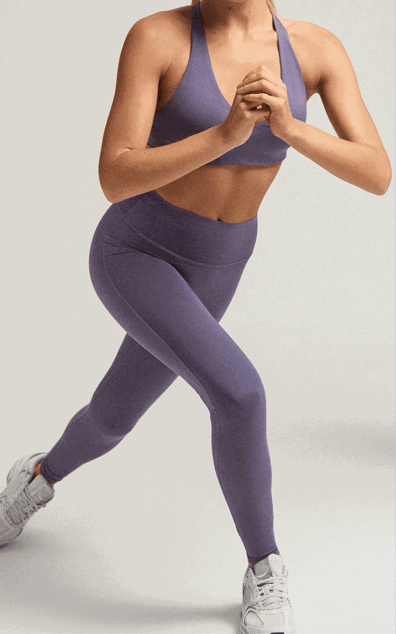 Person in a purple workout outfit and white sneakers performing a lunge exercise against a plain background.