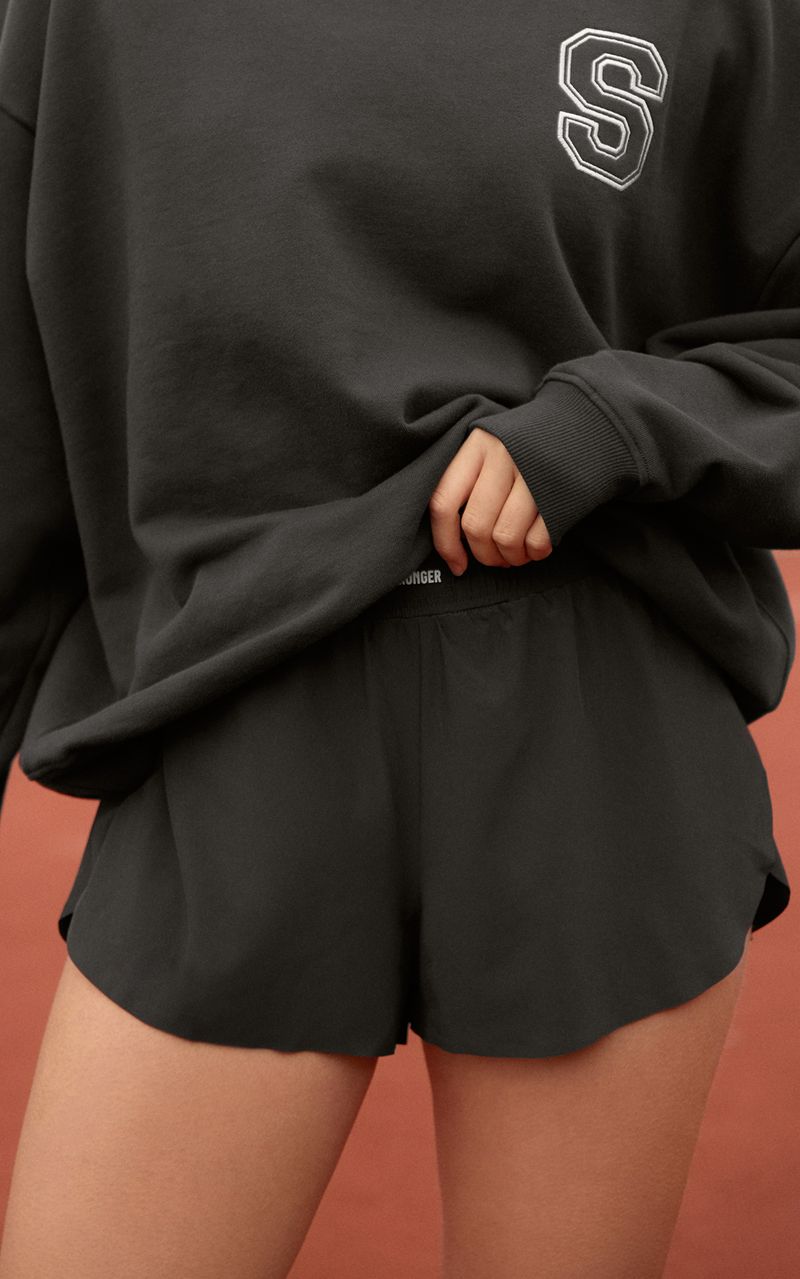 Person wearing a dark sweatshirt with an "S" and matching shorts, standing on a reddish surface, partially tucking the sweatshirt in.