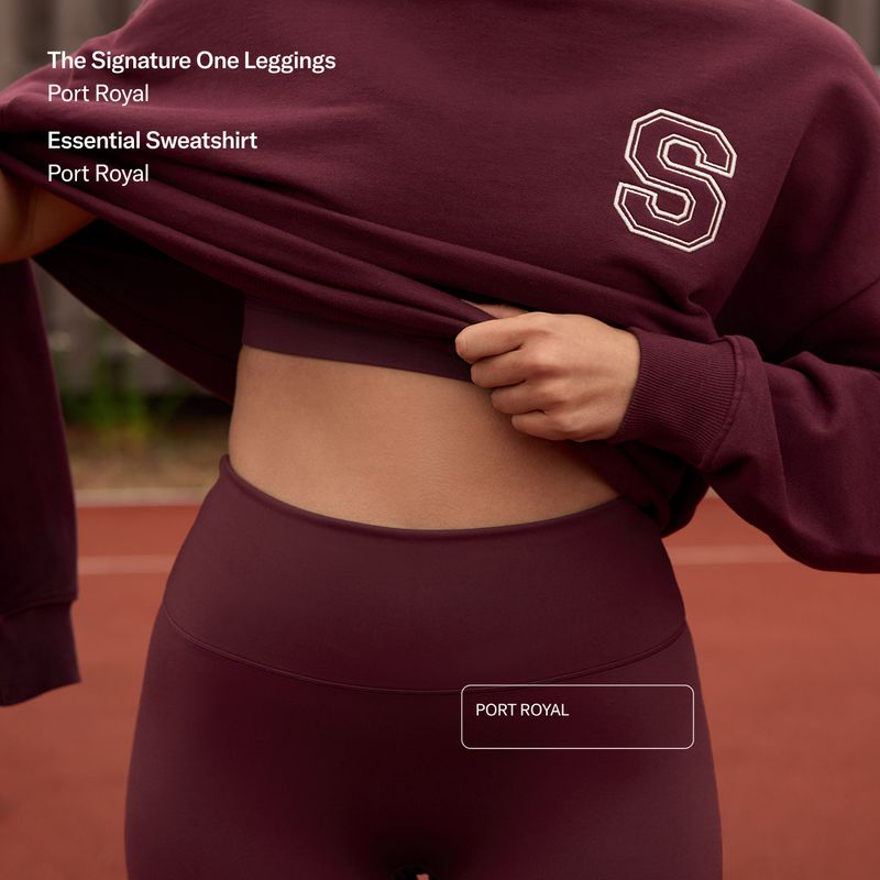 Person lifting a maroon sweatshirt to reveal matching high-waisted leggings. Text displays product names: "The Signature One Leggings" and "Essential Sweatshirt."