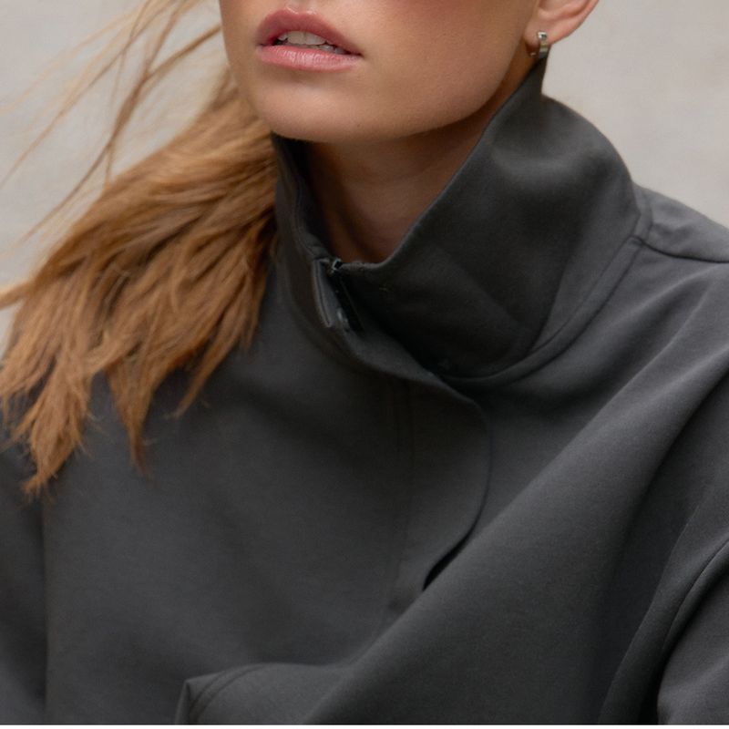 Close-up of a person wearing a high-collared gray jacket, with long hair blowing across the face and small hoop earrings visible.