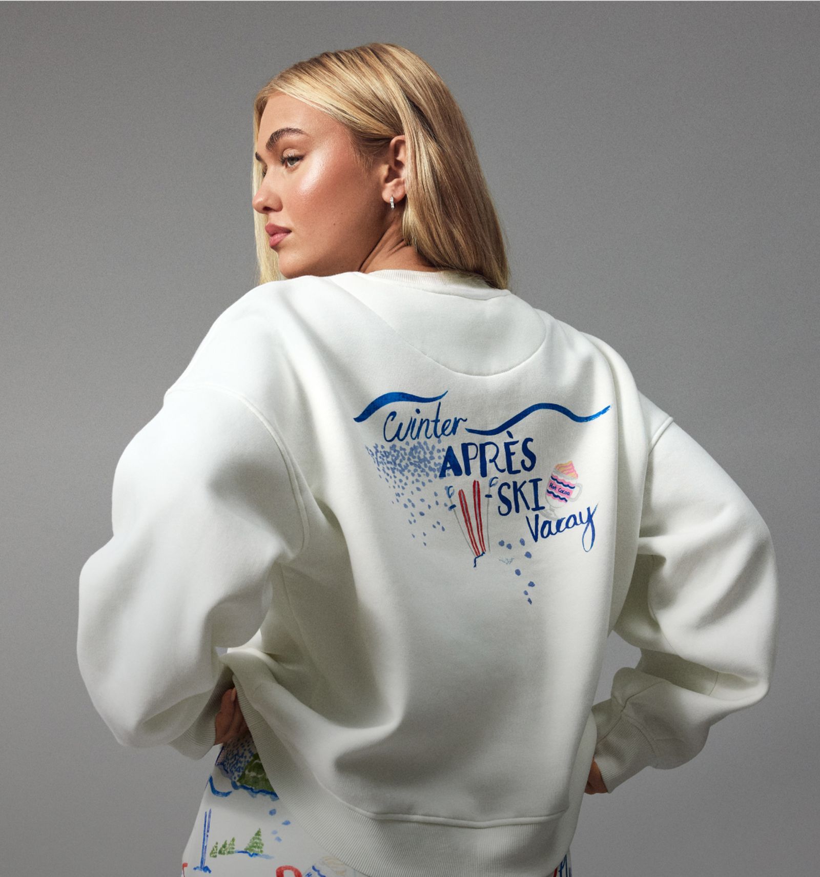 Woman in a white sweatshirt with "Winter Après Ski Vacay" design, featuring blue text and ski graphics, posing against a neutral background.