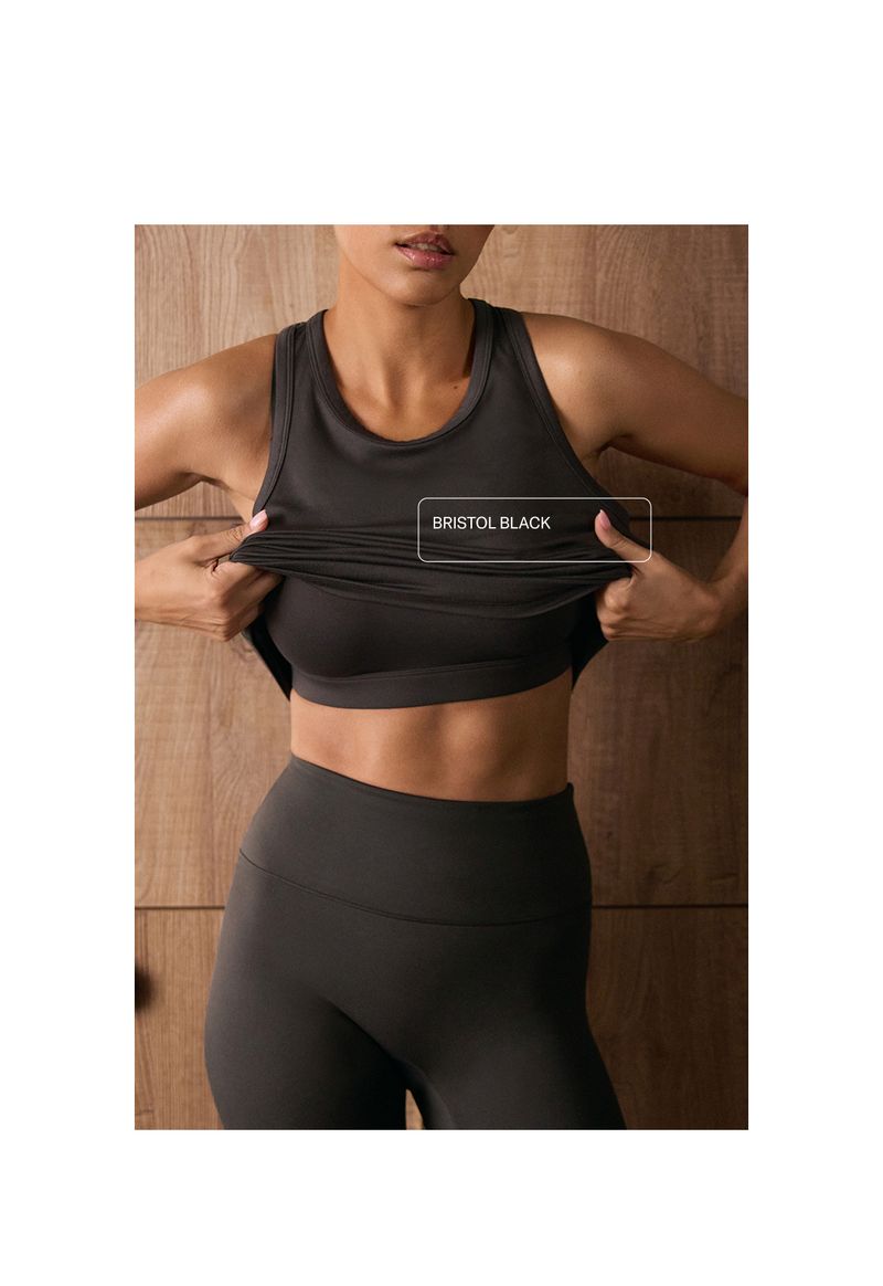 Person wearing a dark sports bra and leggings, lifting a top with "Bristol Black" text, standing against a wooden background.