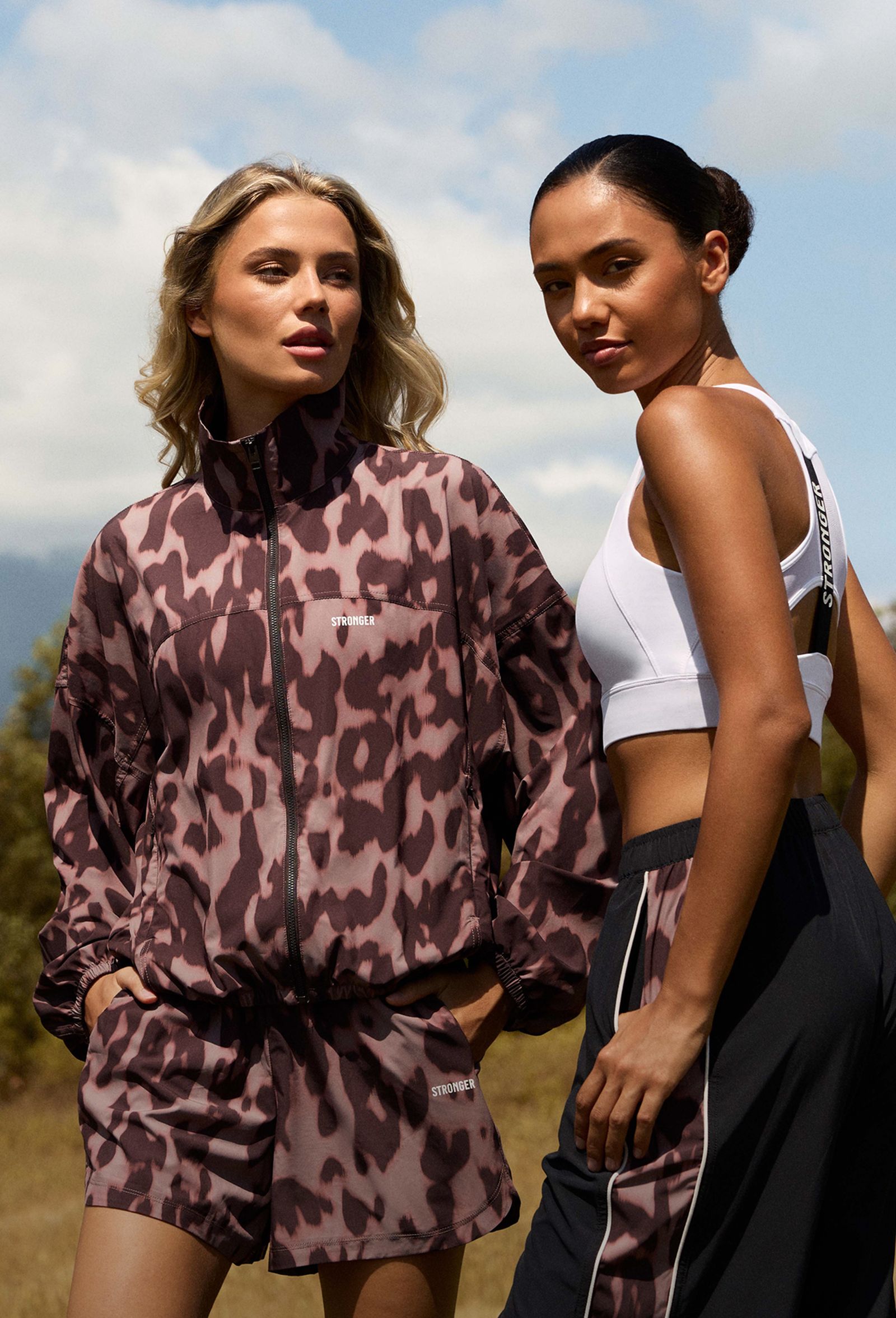 Two women in athletic wear, one in a patterned tracksuit, the other in a white top and dark pants, stand outdoors under a blue sky.