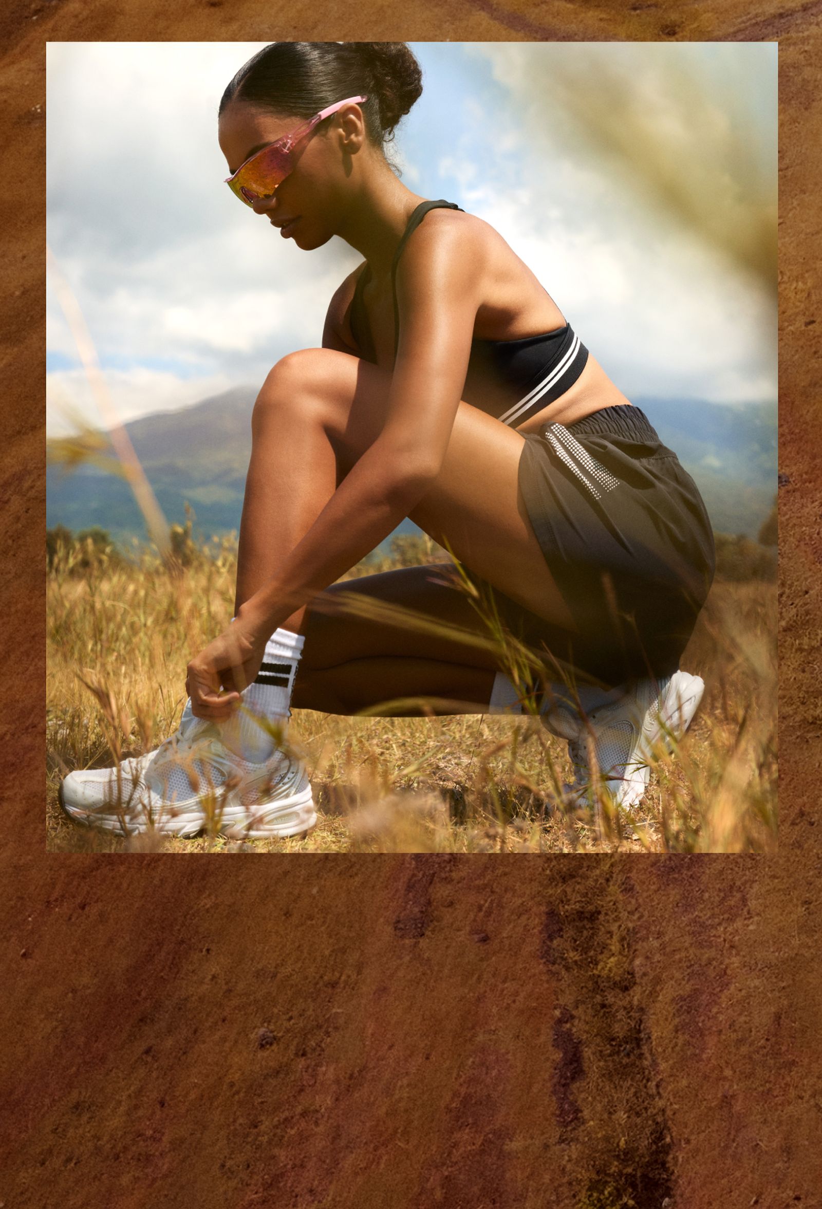 A woman in athletic wear kneels outdoors, adjusting her sneaker, with mountains and sky in the background.