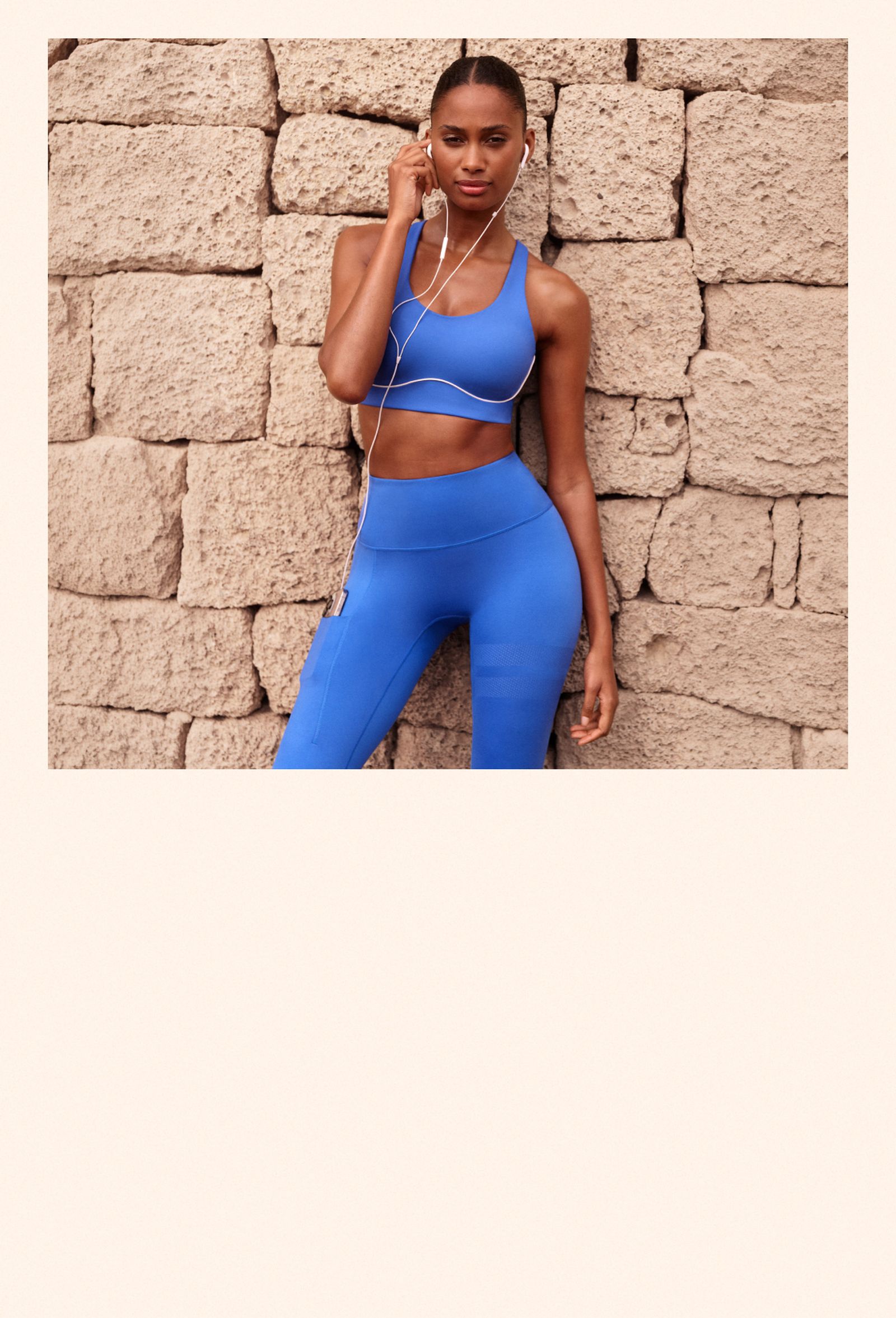 A woman in blue athletic wear stands against a stone wall, listening to music with earphones.