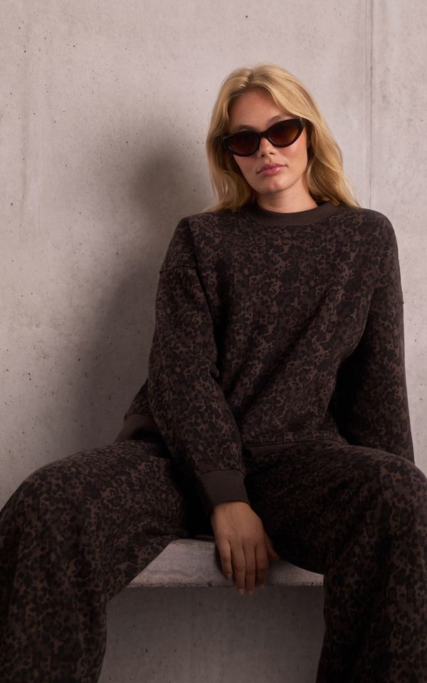 A woman with long blonde hair wearing dark sunglasses and a leopard print sweatshirt sits against a concrete wall.