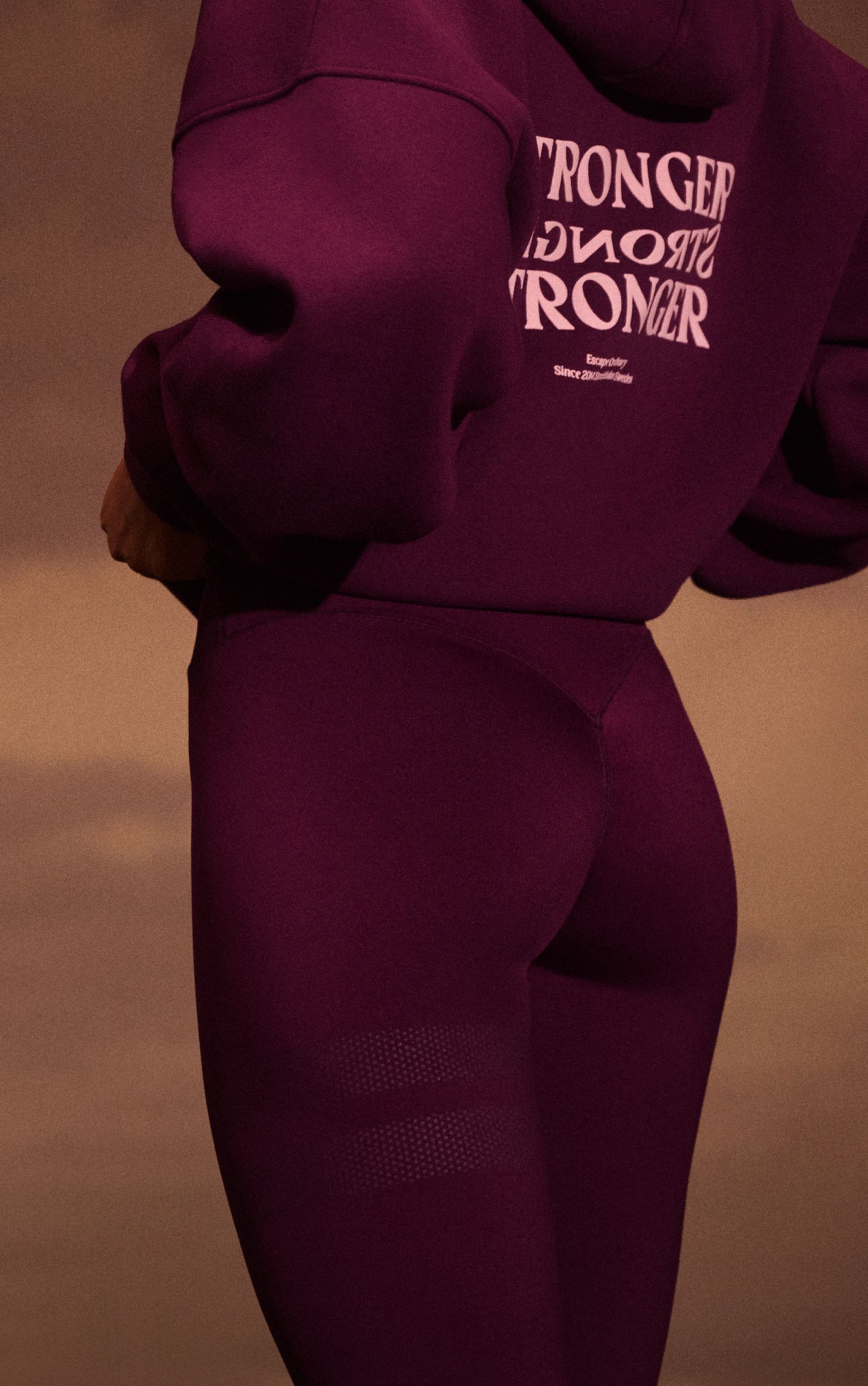 Person wearing a purple hoodie with "STRONGER" text and matching leggings, viewed from the side, against a warm background.
