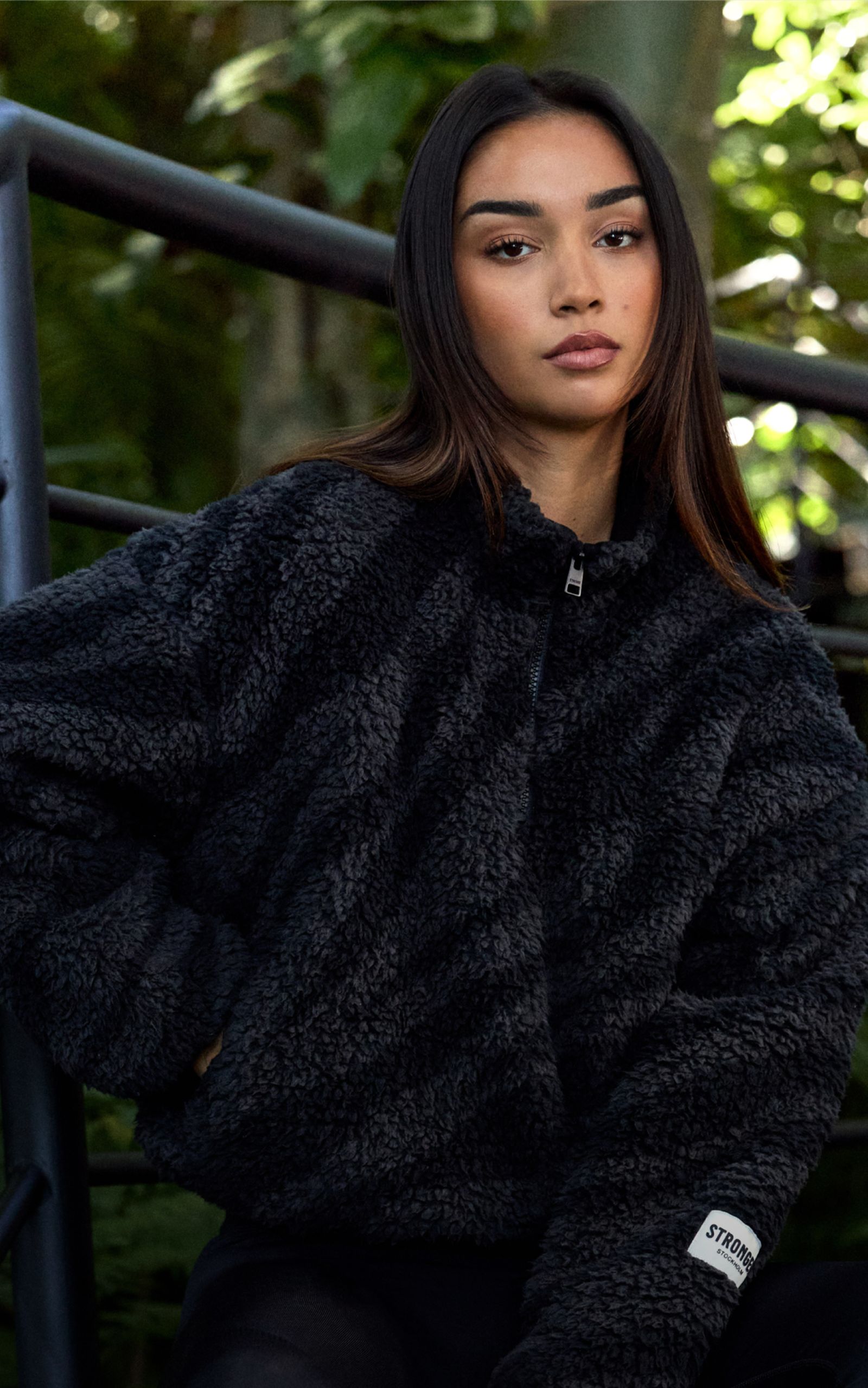 A person with long dark hair wearing a fuzzy black jacket sits outdoors, with a leafy background and a railing beside them.