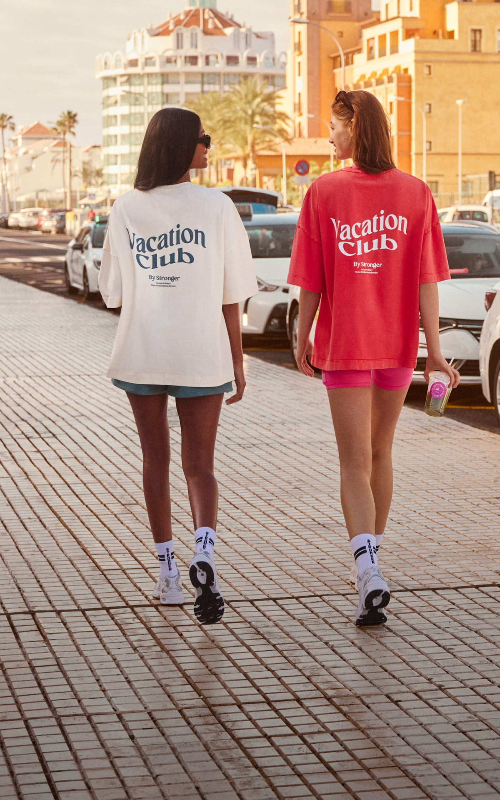 Two people walking on a sunlit boardwalk, wearing t-shirts with "Vacation Club" printed on the back. Cars and buildings line the background.