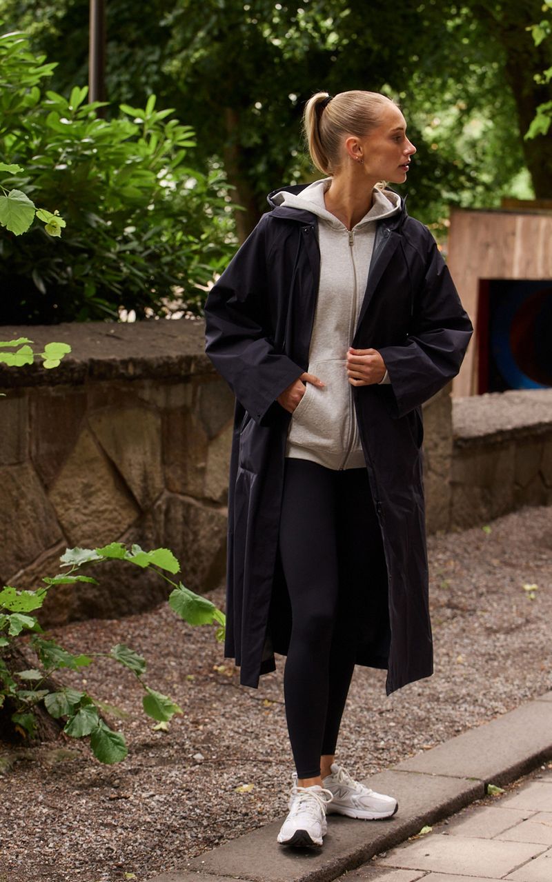 Woman in a long black coat over a gray hoodie and black leggings, walking on a path surrounded by greenery.