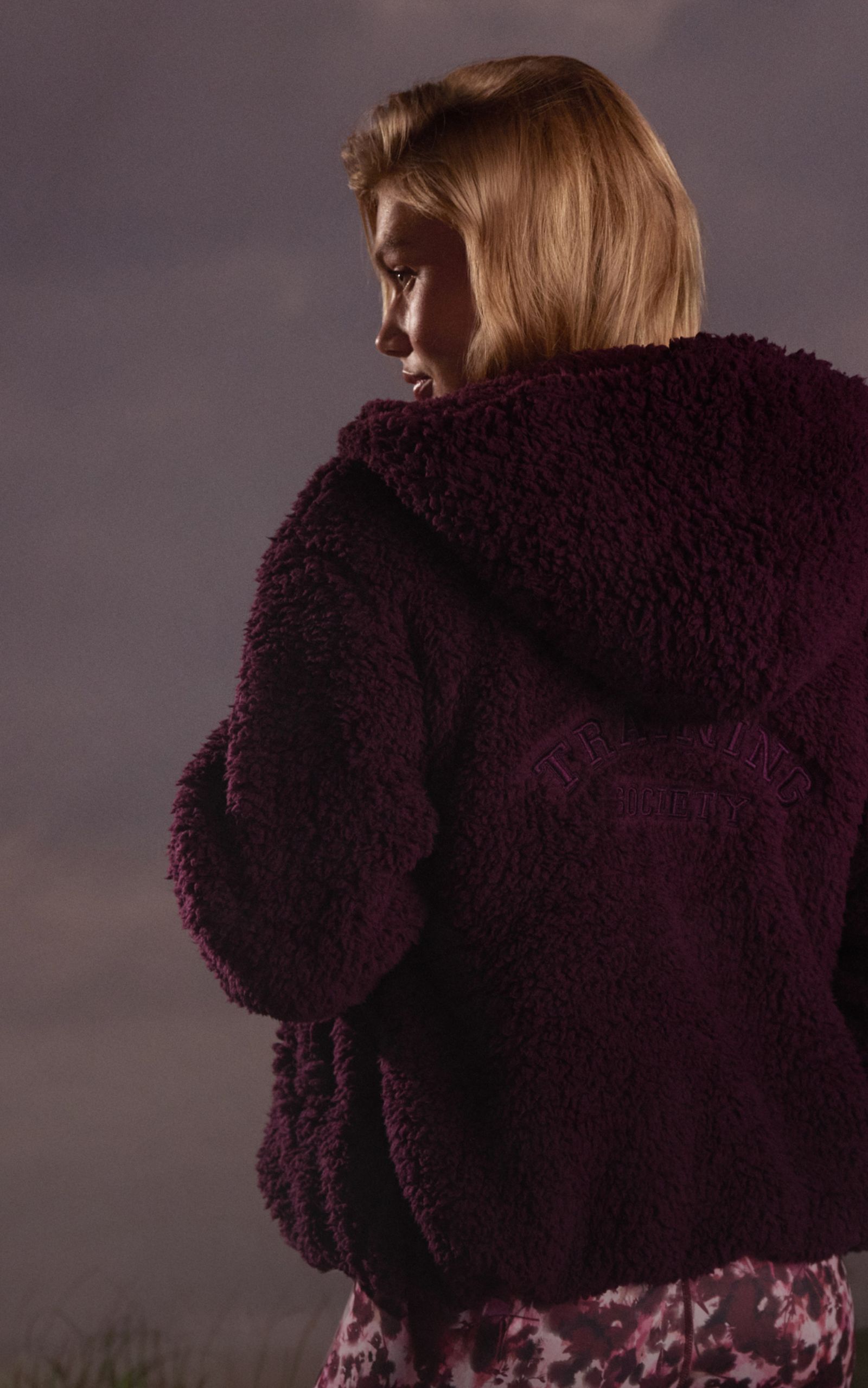 A person with short blond hair wearing a dark purple, textured hoodie with "Training Society" text on the back, and a floral garment underneath.