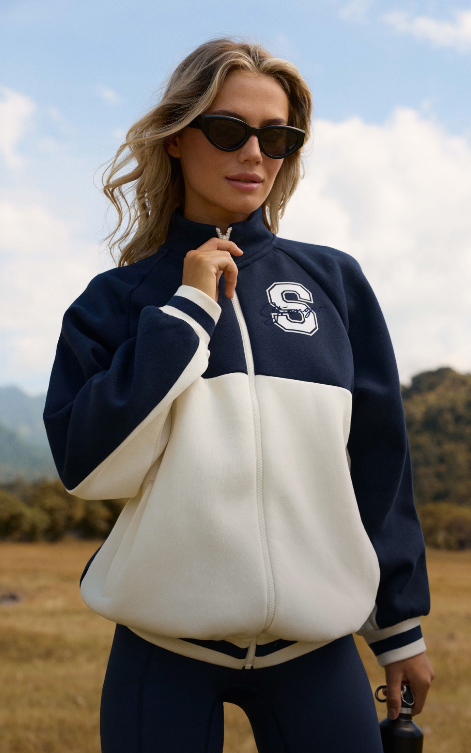 A person wearing sunglasses and a navy and white jacket with "S" logo stands outdoors against a blurred natural background.