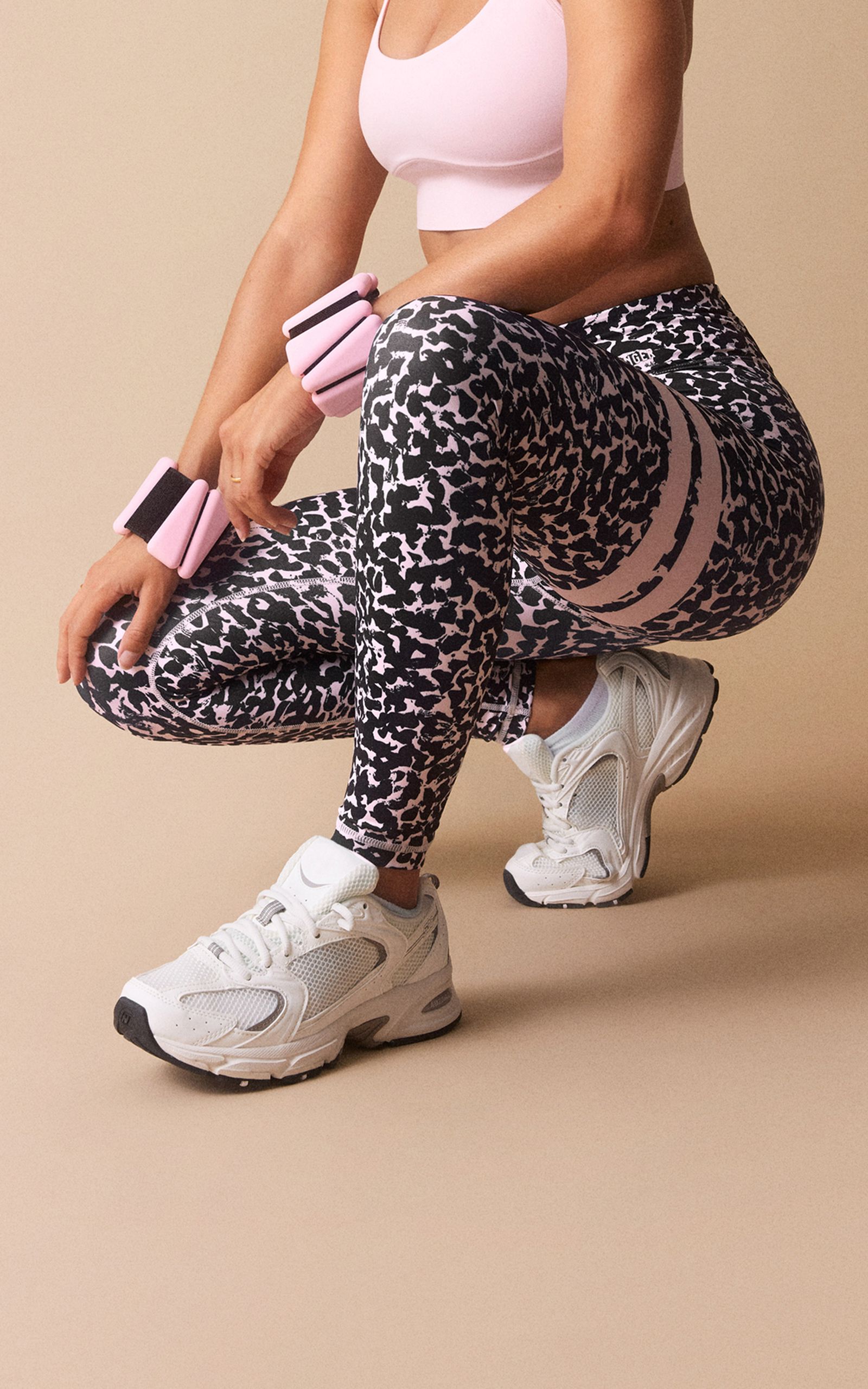 Person in pink sports bra, patterned leggings, and white sneakers, crouching with pink wrist weights, against beige background.