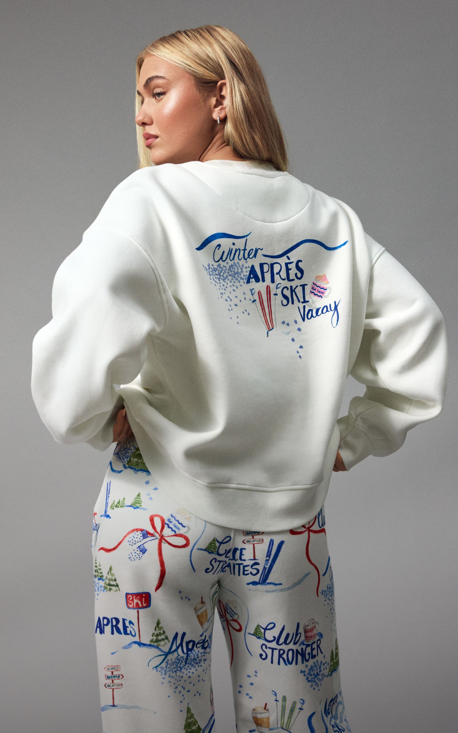 Woman wearing a white sweatshirt with "Winter Après Ski Vacay" text, paired with colorful printed pants featuring ski-themed graphics.