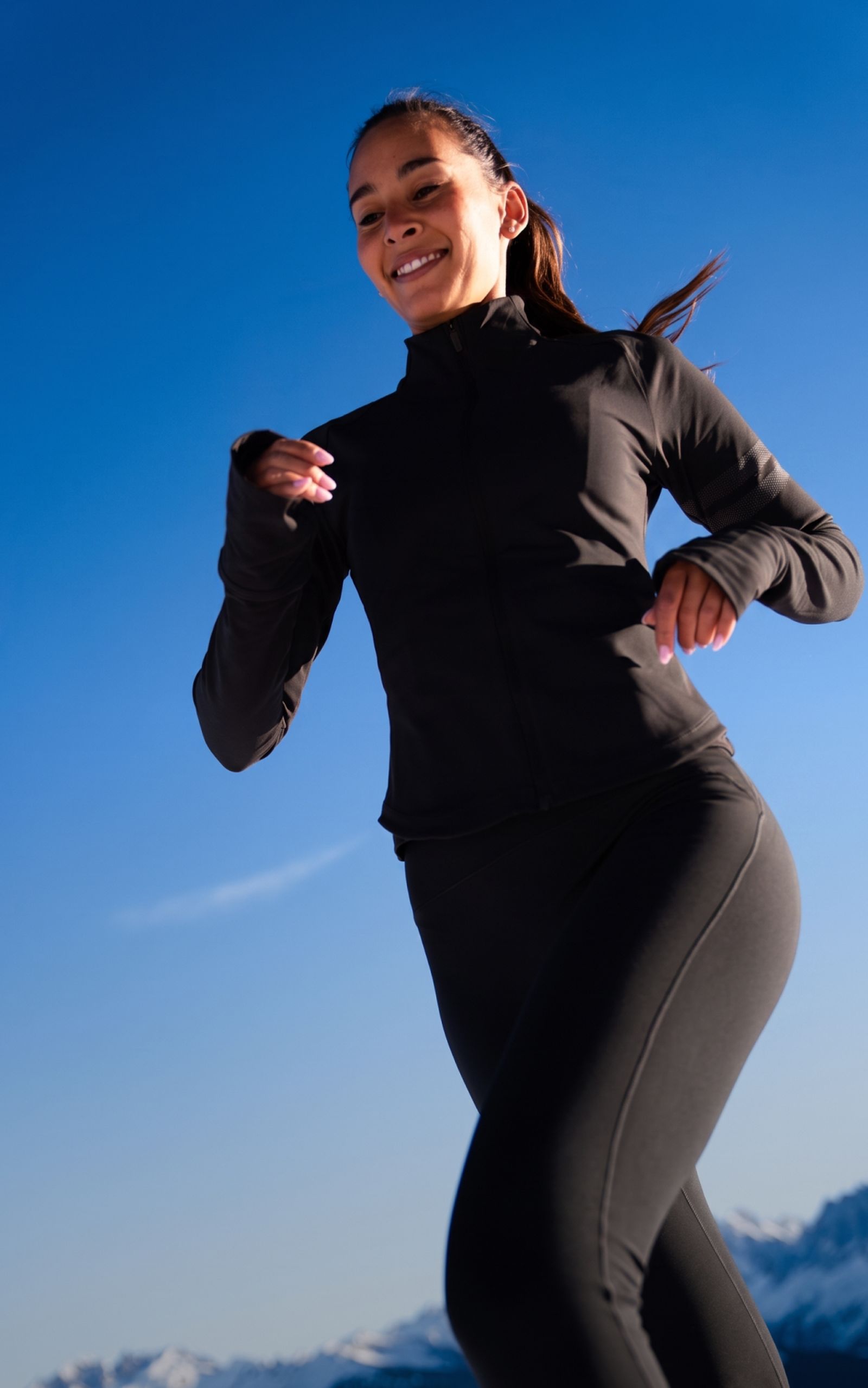 A woman in athletic wear jogs outdoors against a clear blue sky, with snowy mountains in the background.