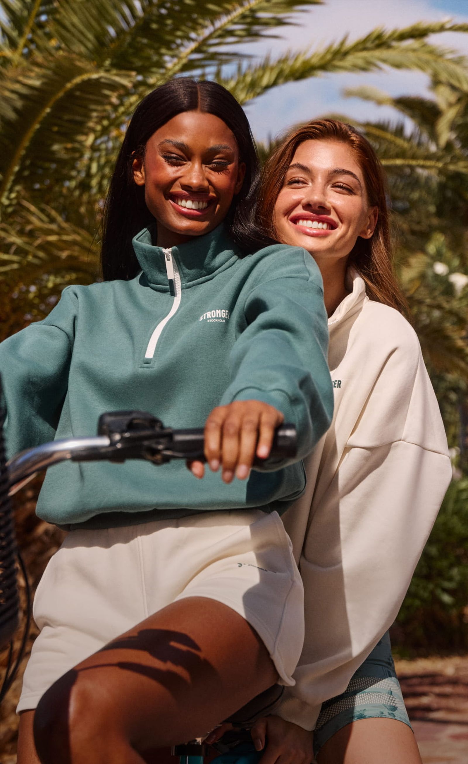 Two women smiling while riding a bicycle together. They are wearing casual sweatshirts and enjoying a sunny day with palm trees in the background.