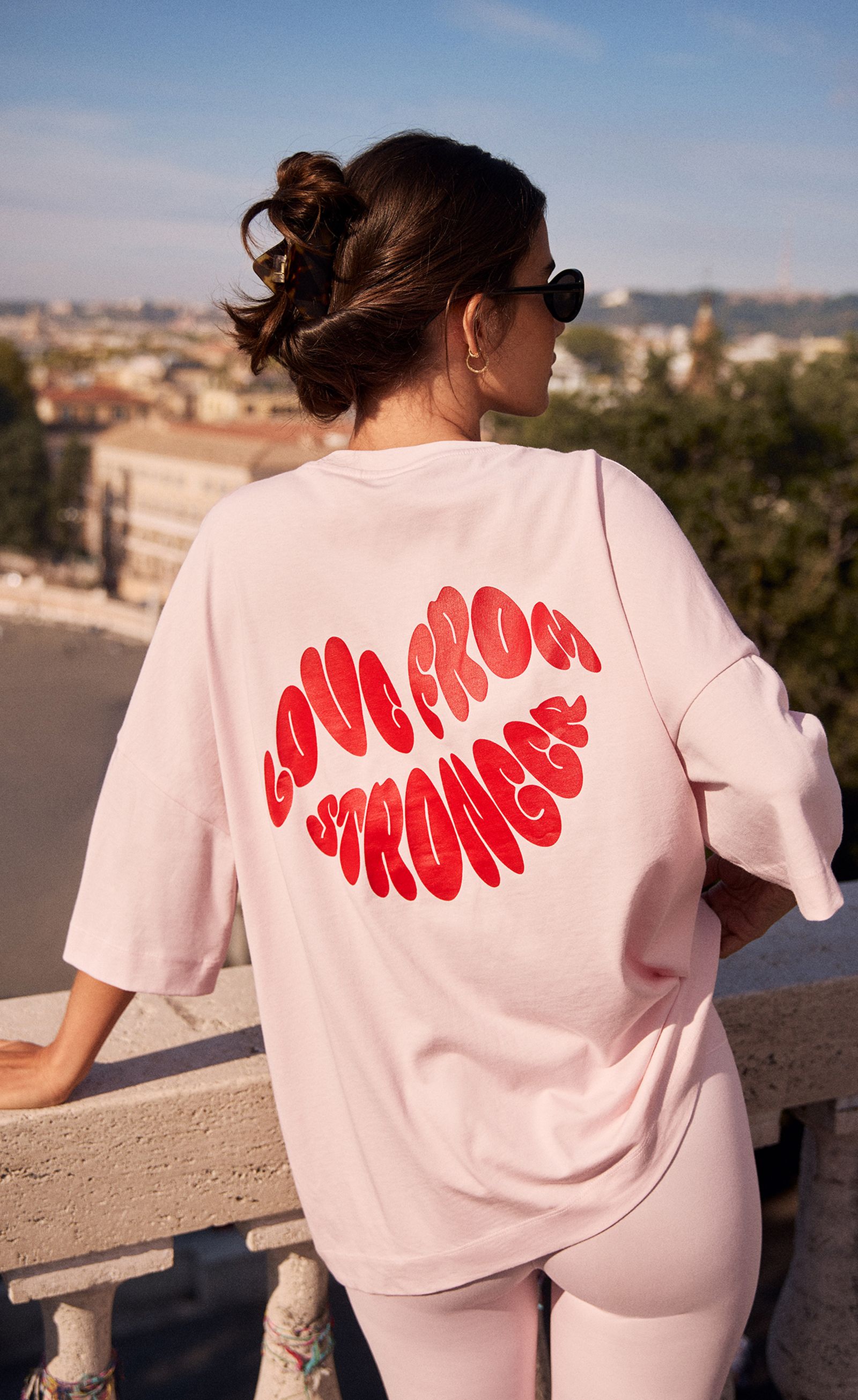 Woman in sunglasses and pink outfit with "LOVE FROM STRONGER" on her shirt, leaning on a balcony, overlooking a cityscape.