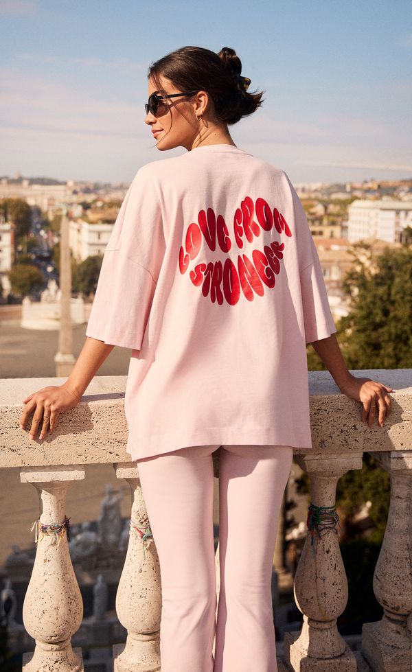 Woman in pink outfit, sunglasses, facing city skyline on a balcony. Shirt reads "Love from Roma." Bright and sunny day.