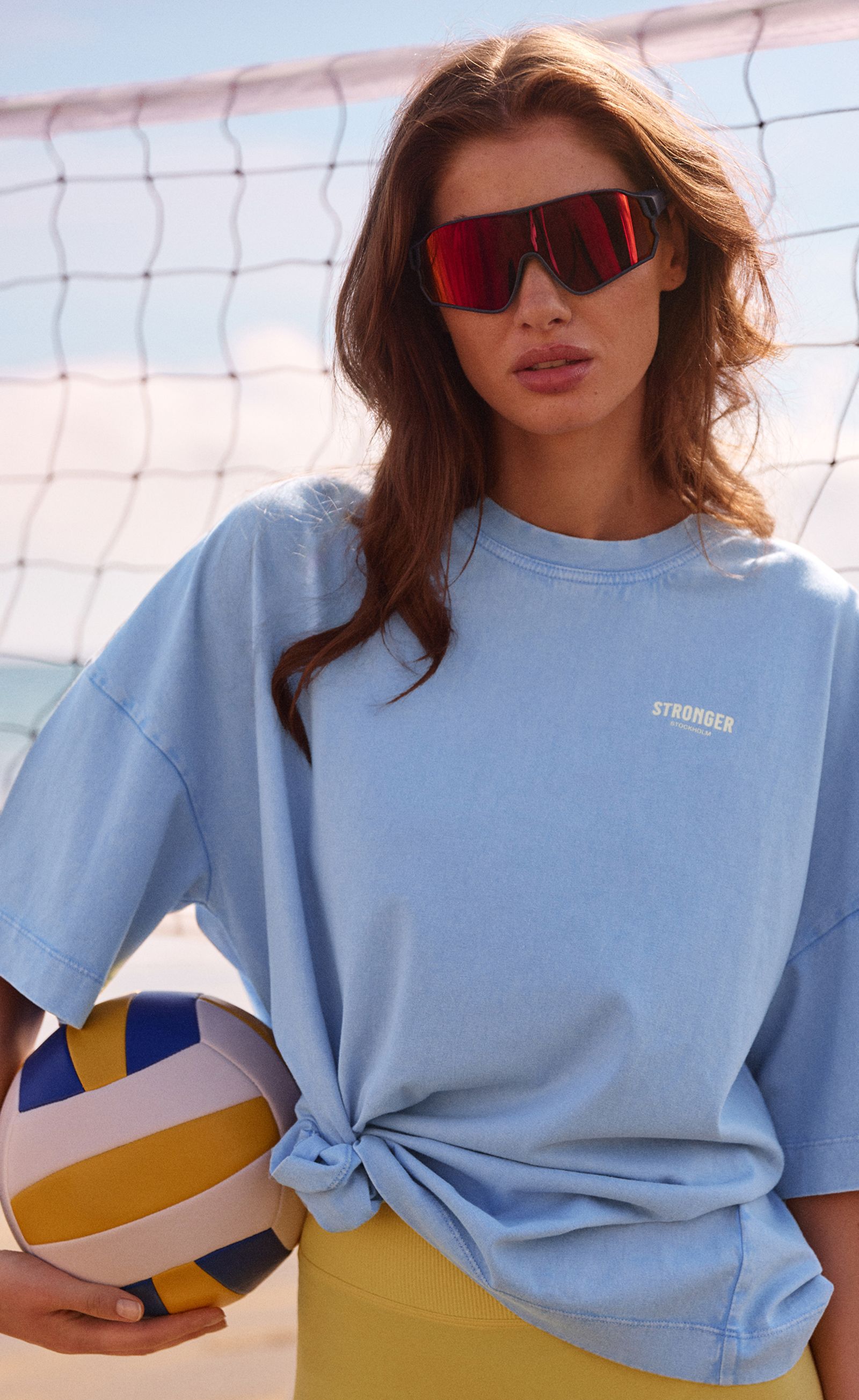 Woman in a blue tee and red sunglasses holds a volleyball on a sandy beach with a net in the background, under a clear sky.