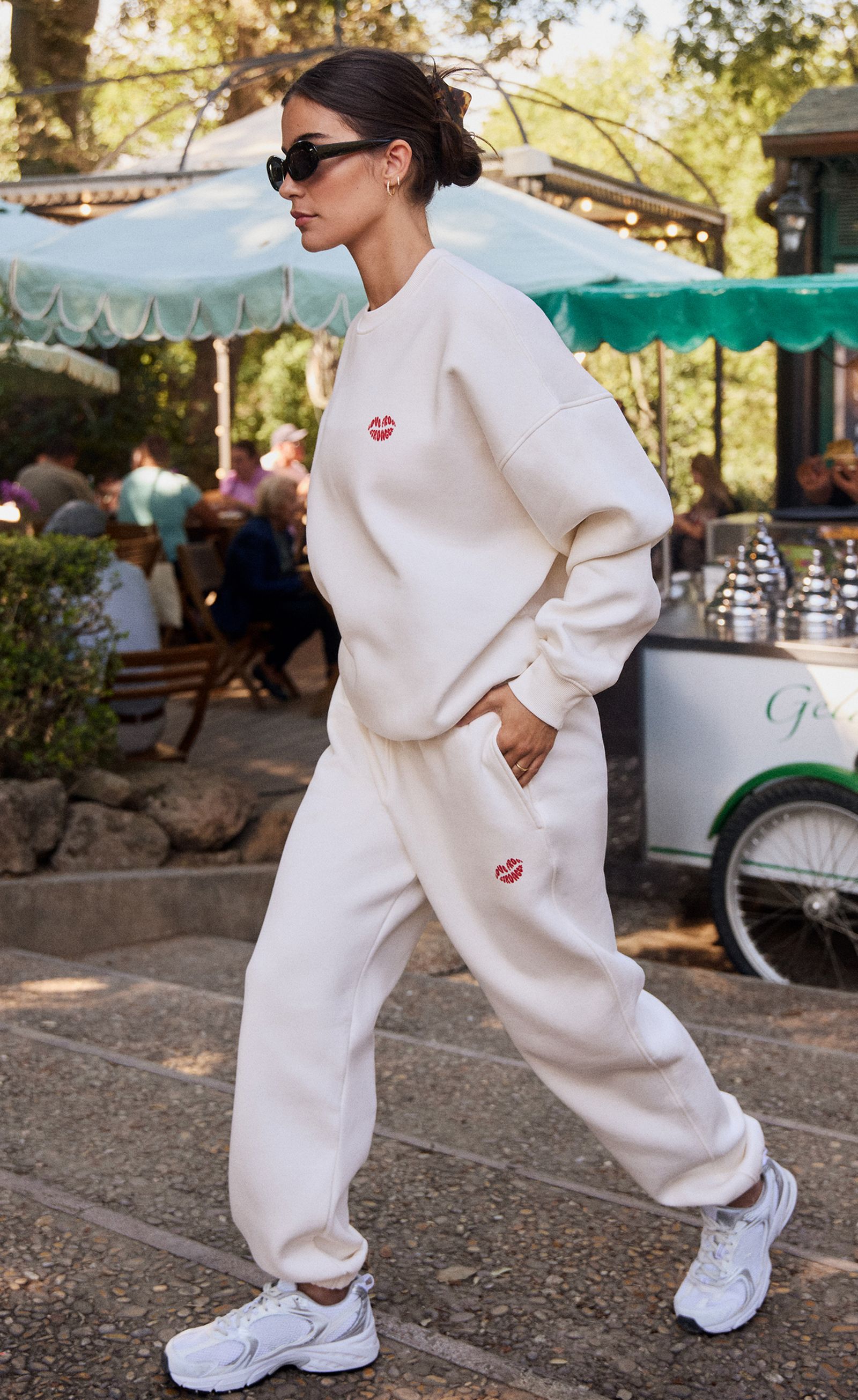 Woman in a cream sweatsuit and sunglasses walking outdoors near a cafe, with trees and people in the background.