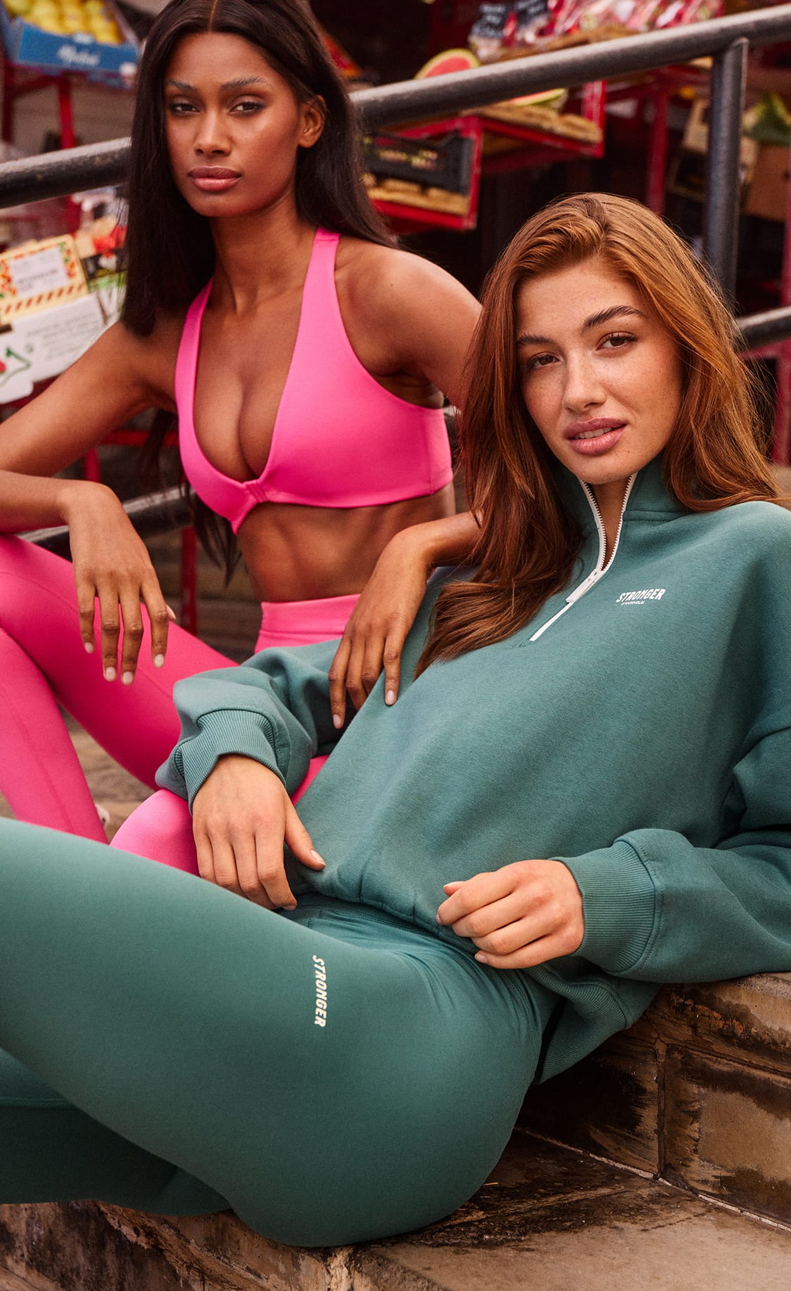 Two women in stylish activewear; one in a pink sports outfit, the other in a green tracksuit, sitting outdoors near stairs.