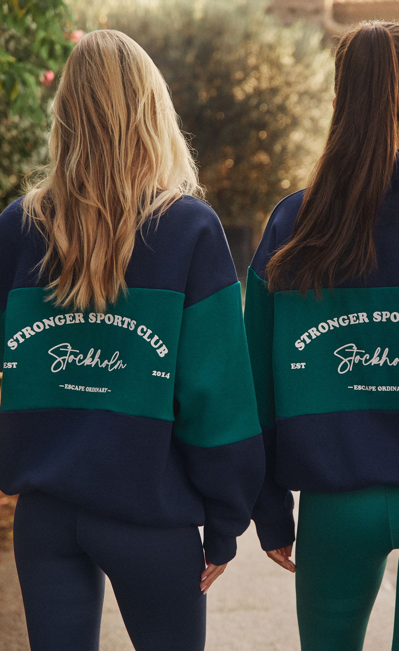 Two people standing with backs to camera, wearing navy-and-green "Stronger Sports Club Stockholm" sweatshirts.