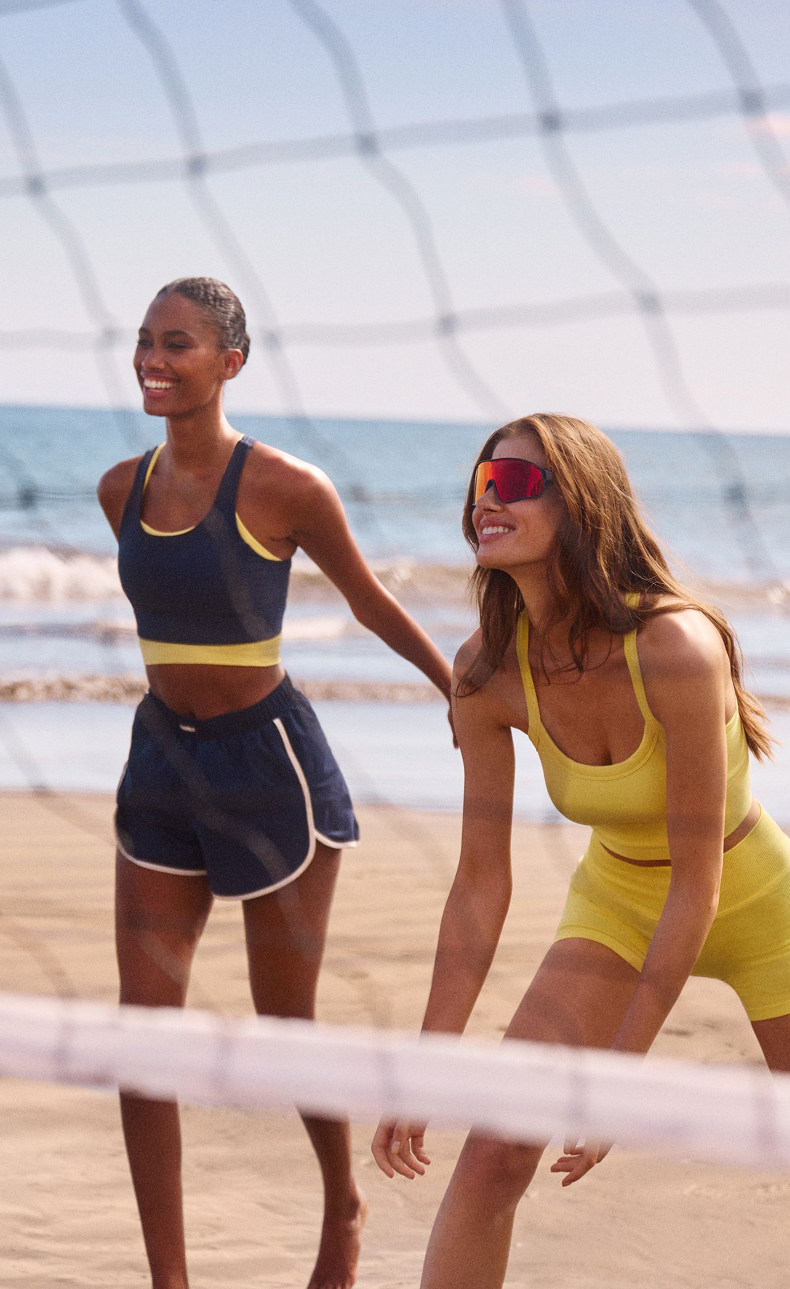 Two women in athletic outfits playing beach volleyball, with the net visible and the sea in the background.
