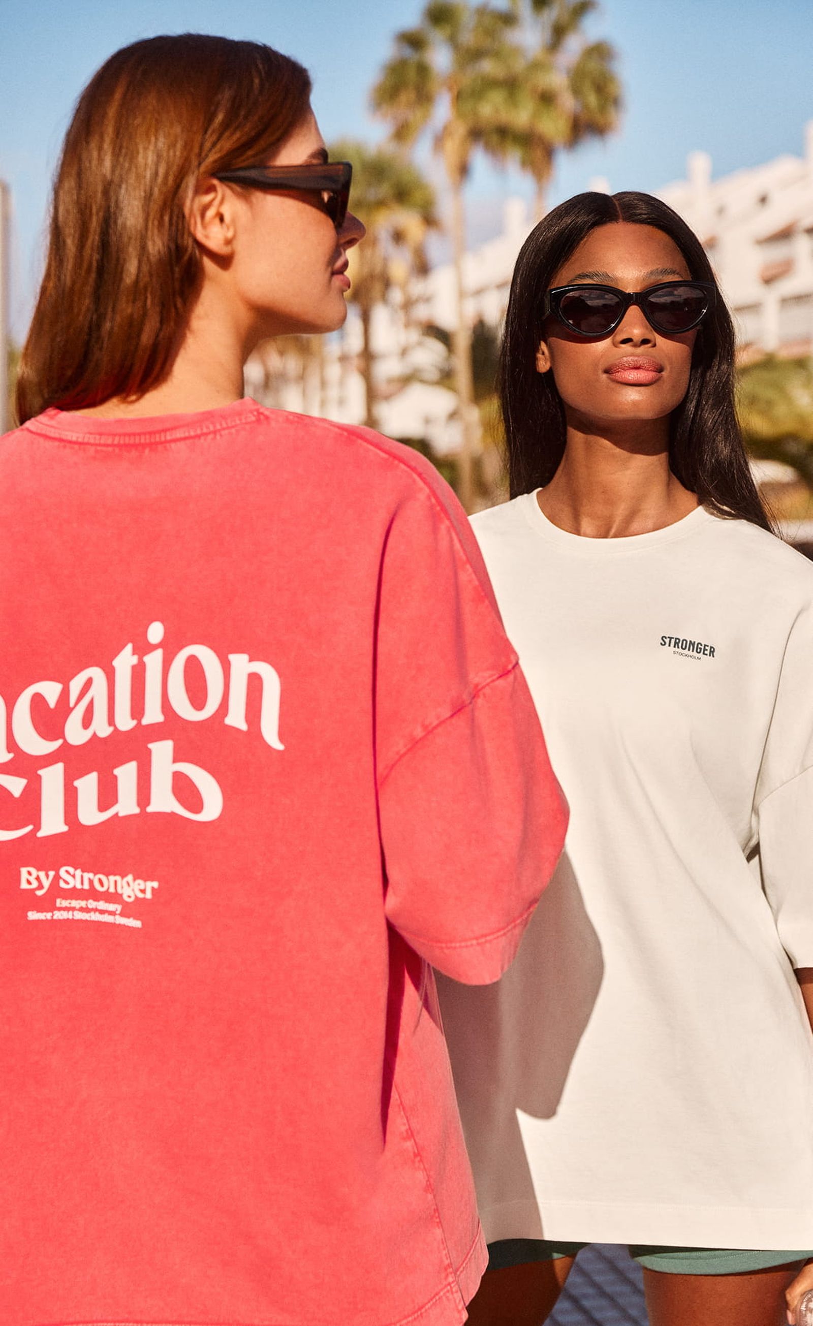 Two women wearing sunglasses and oversized T-shirts, one red with "Vacation Club," outdoors with palm trees and white buildings in the background.