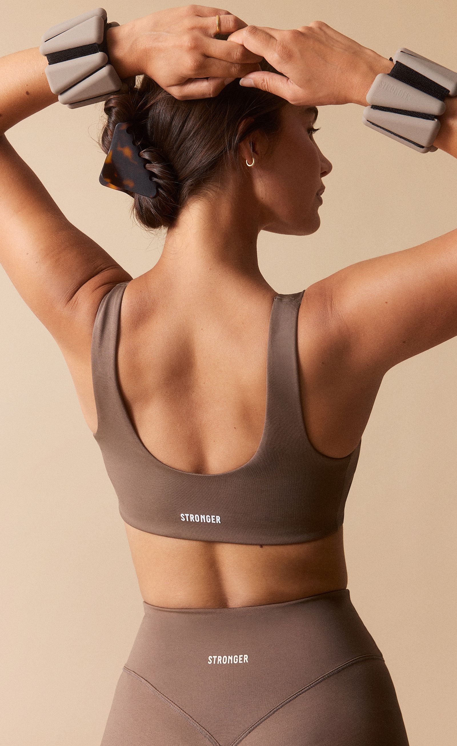 Woman in brown athletic wear with arm weights, seen from behind, hair in a clip, arms raised and hands on head against a beige background.