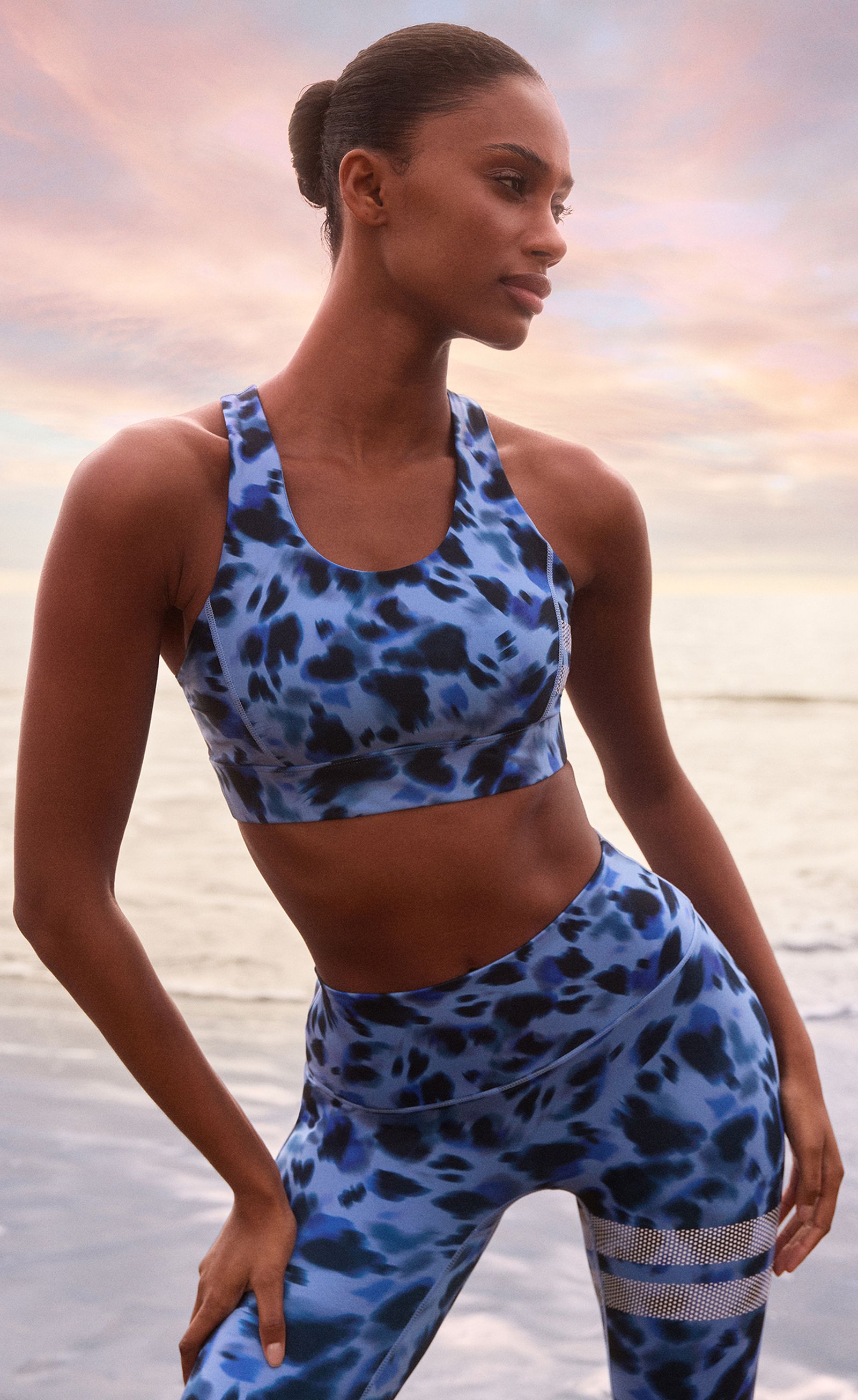 Woman in blue leopard-print activewear poses by the ocean under a colorful sunset sky.