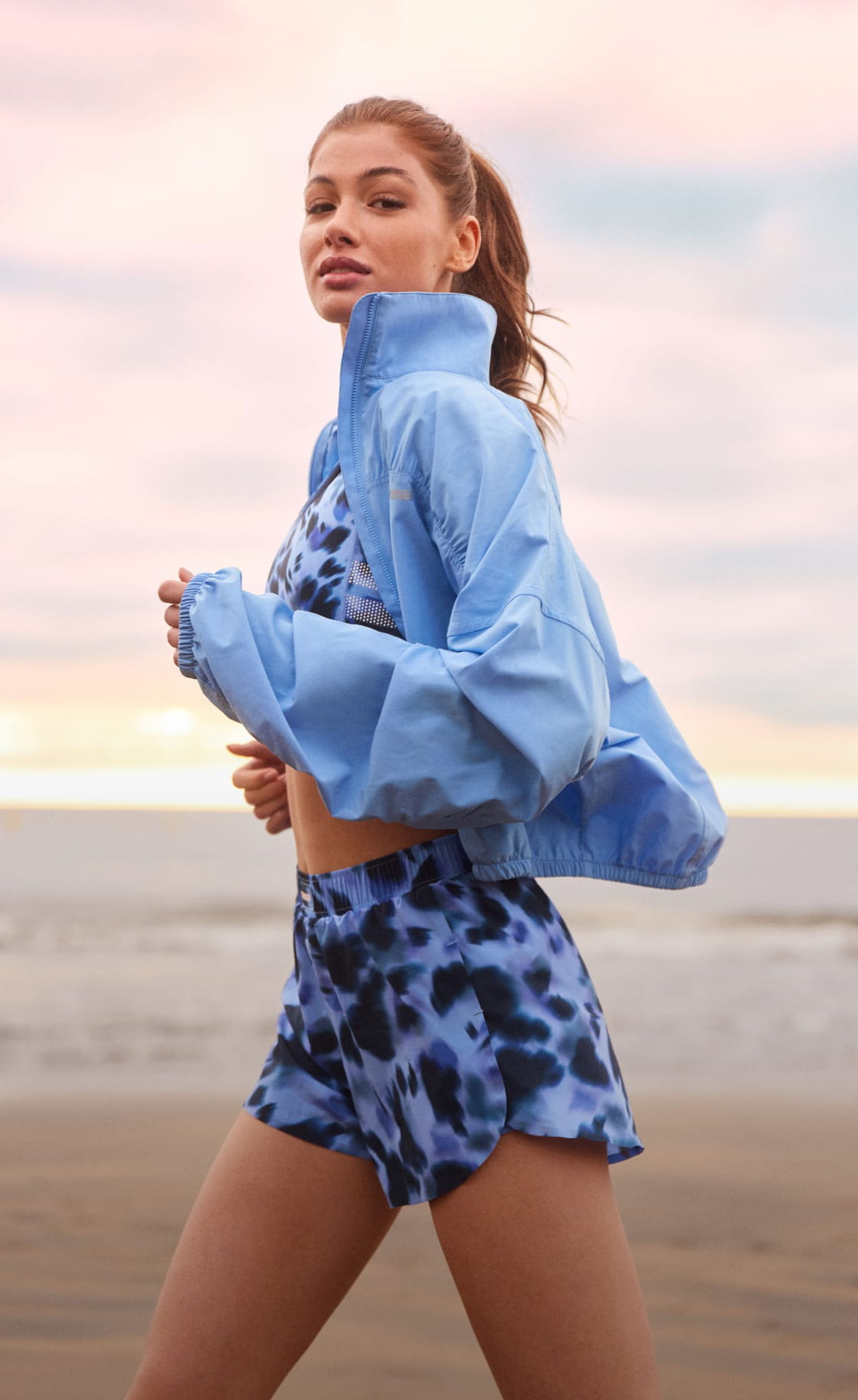 A woman in a blue athletic outfit jogs along a beach at sunrise, with a pink and blue sky in the background.