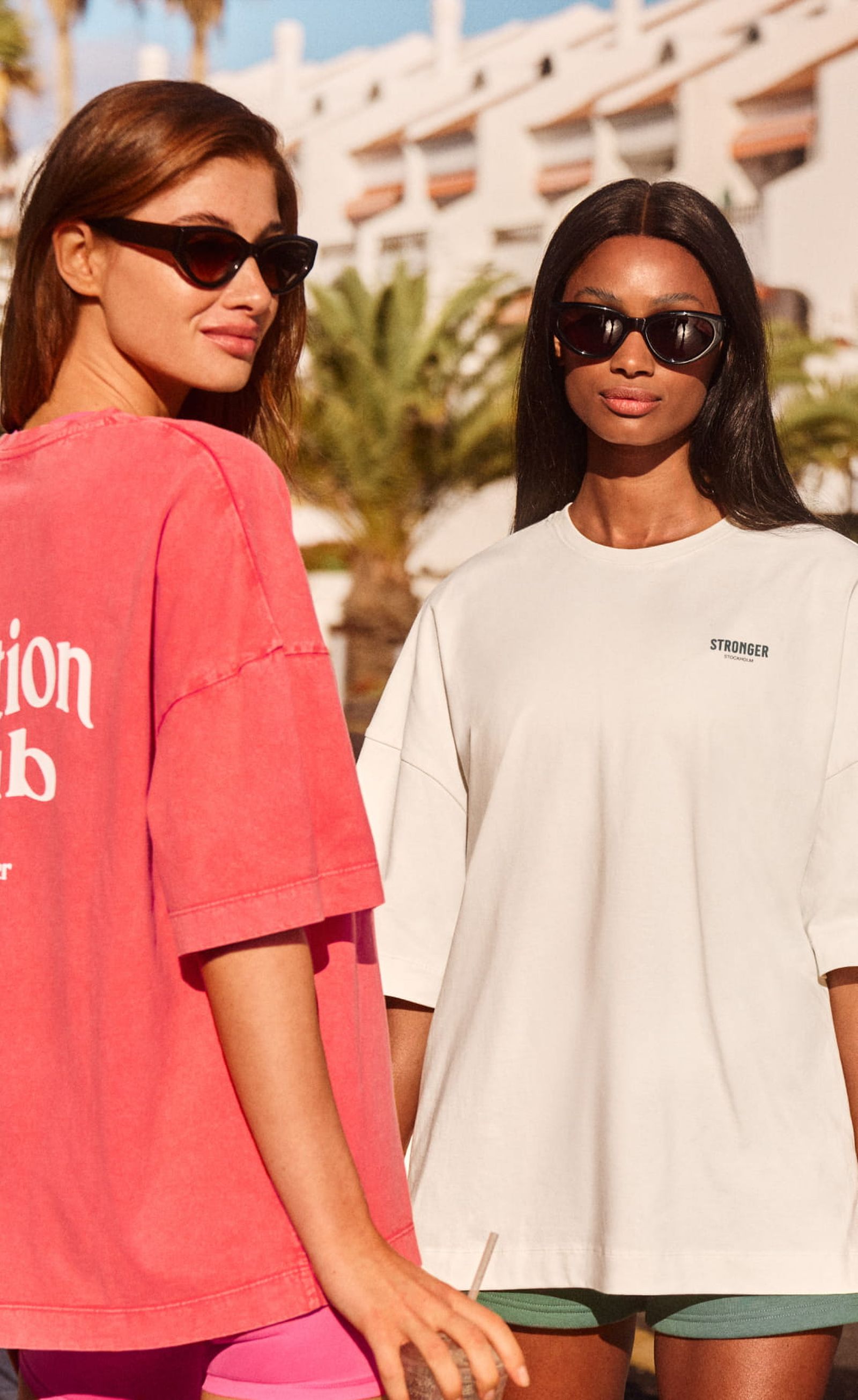 Two women in sunglasses and casual t-shirts stand outdoors, one wearing red and the other white, with palm trees and a building in the background.
